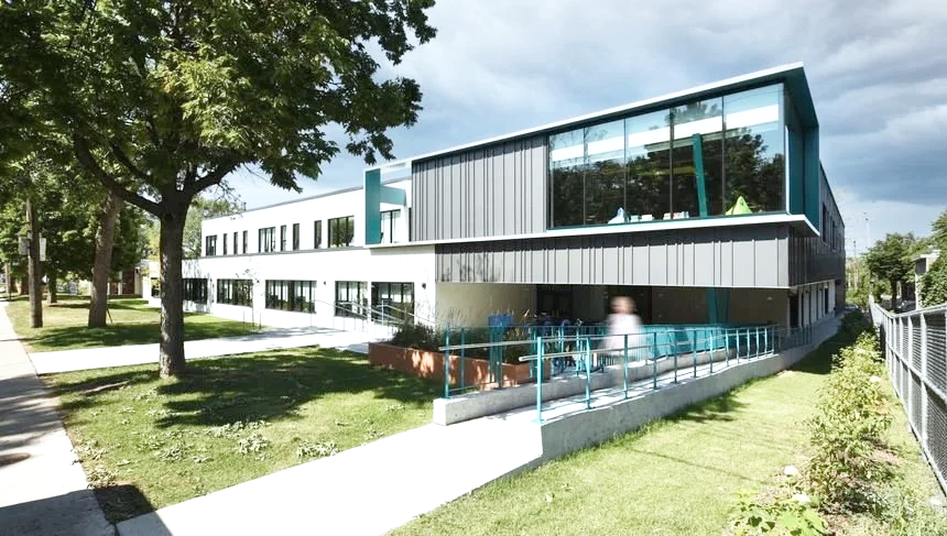 Modern school building with large glass windows, surrounded by green trees and lawn, under a cloudy sky.