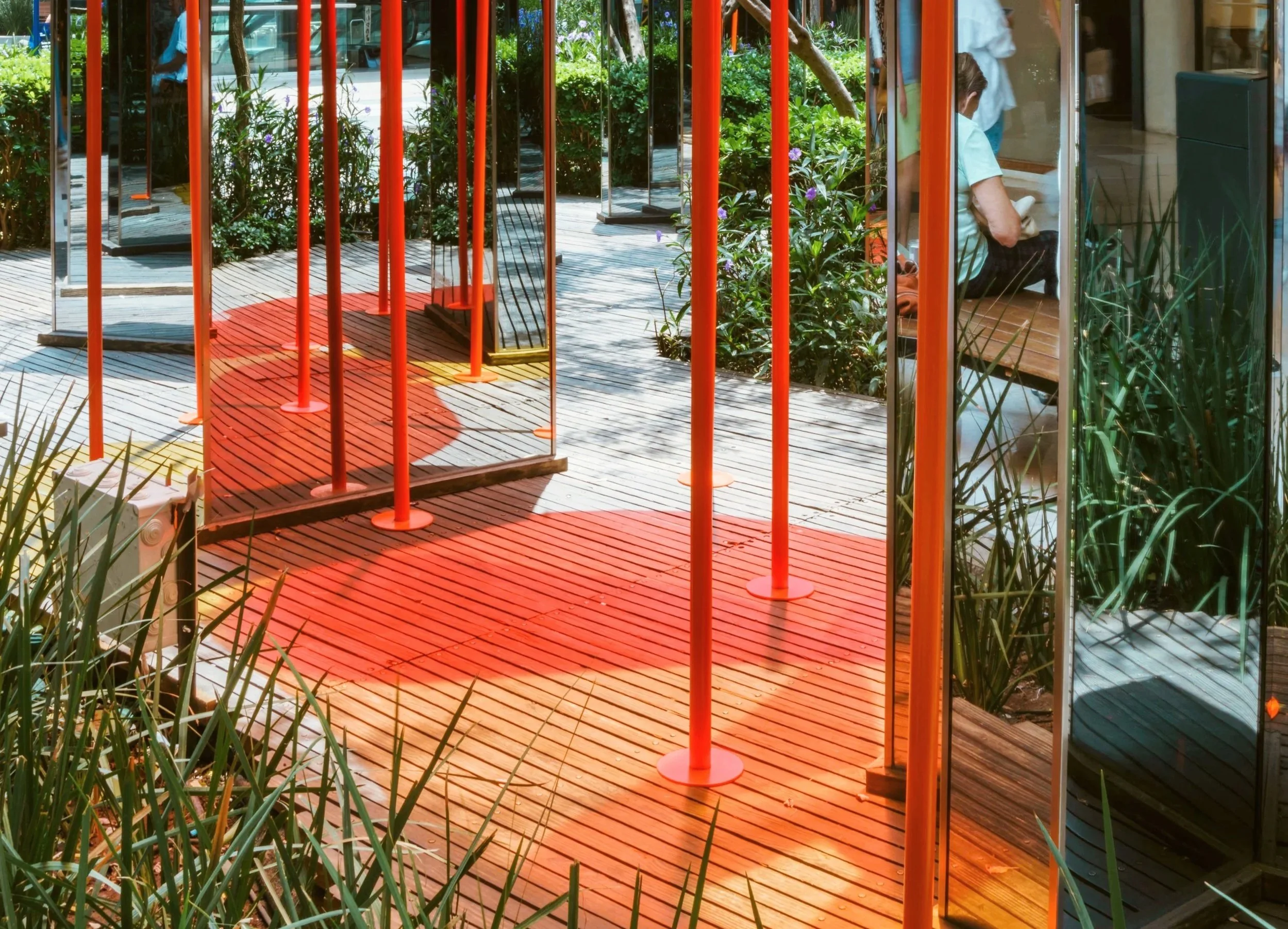 A glass door leads to a small outdoor area with a wooden deck, orange metal posts, and lush green plants with purple flowers. Inside, there is a person sitting on a bench, possibly reading or using a device, near the window.