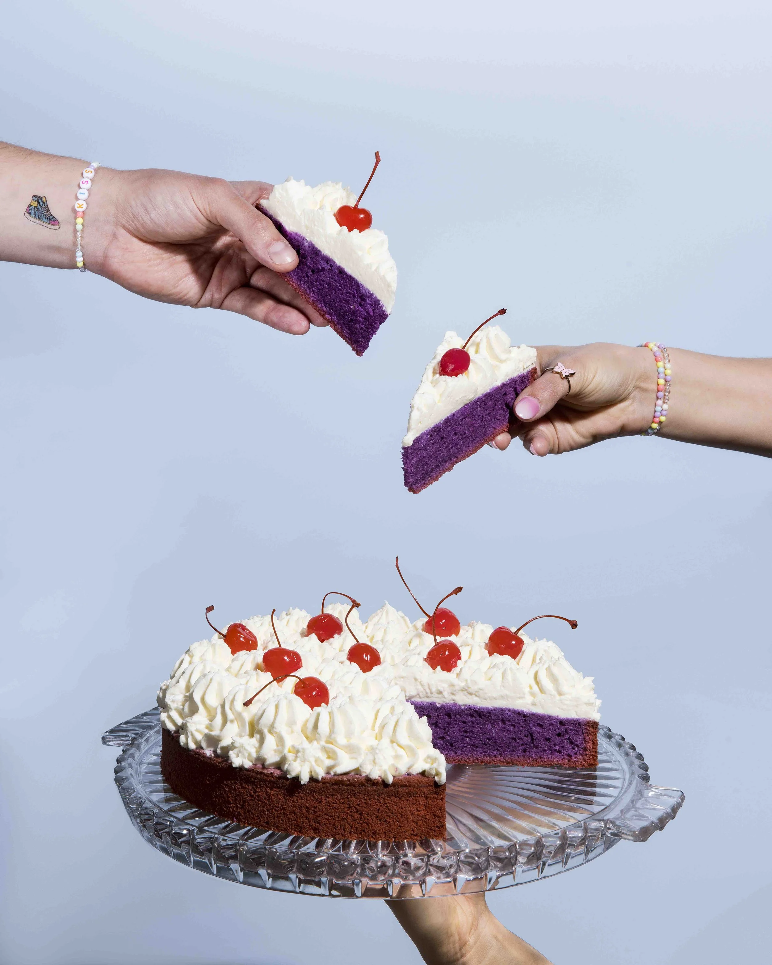 Eine Hand hält eine große Torte mit lila Boden, weißer Sahne und Kirschen auf einem Kristall-Servierteller, während eine andere Hand ein Stück Torte hochhebt. Die Torte hat eine cremige Oberseite mit Sahne und Kirschen als Dekoration.