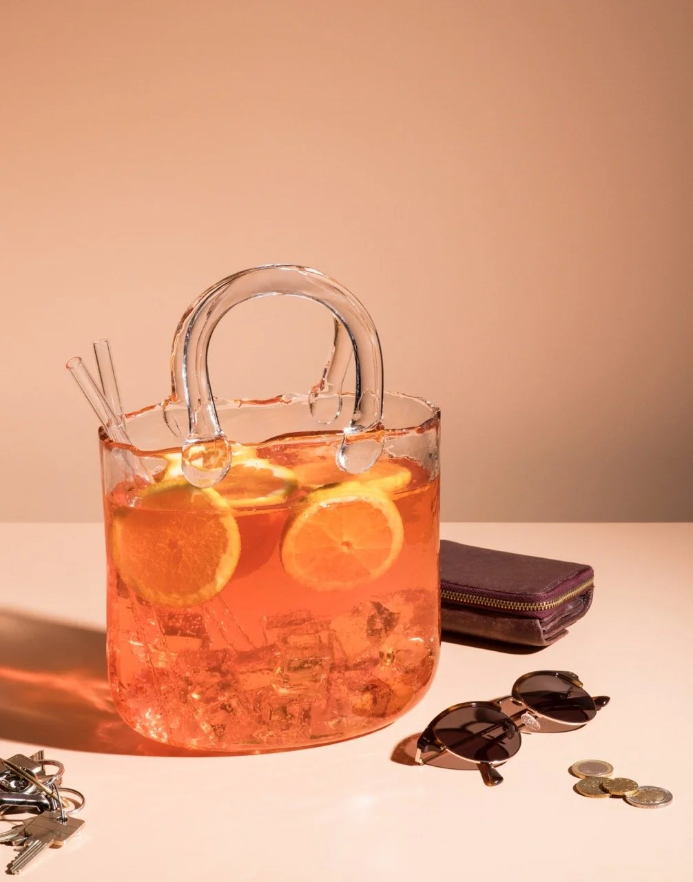 Auf dem Bild ist eine große Glas-Kanne mit Orangensaft und Orangenscheiben, umgeben von Sonnenbrillen, Geldscheinen, einem Geldbeutel und einem Schlüssel bei warmem Licht.