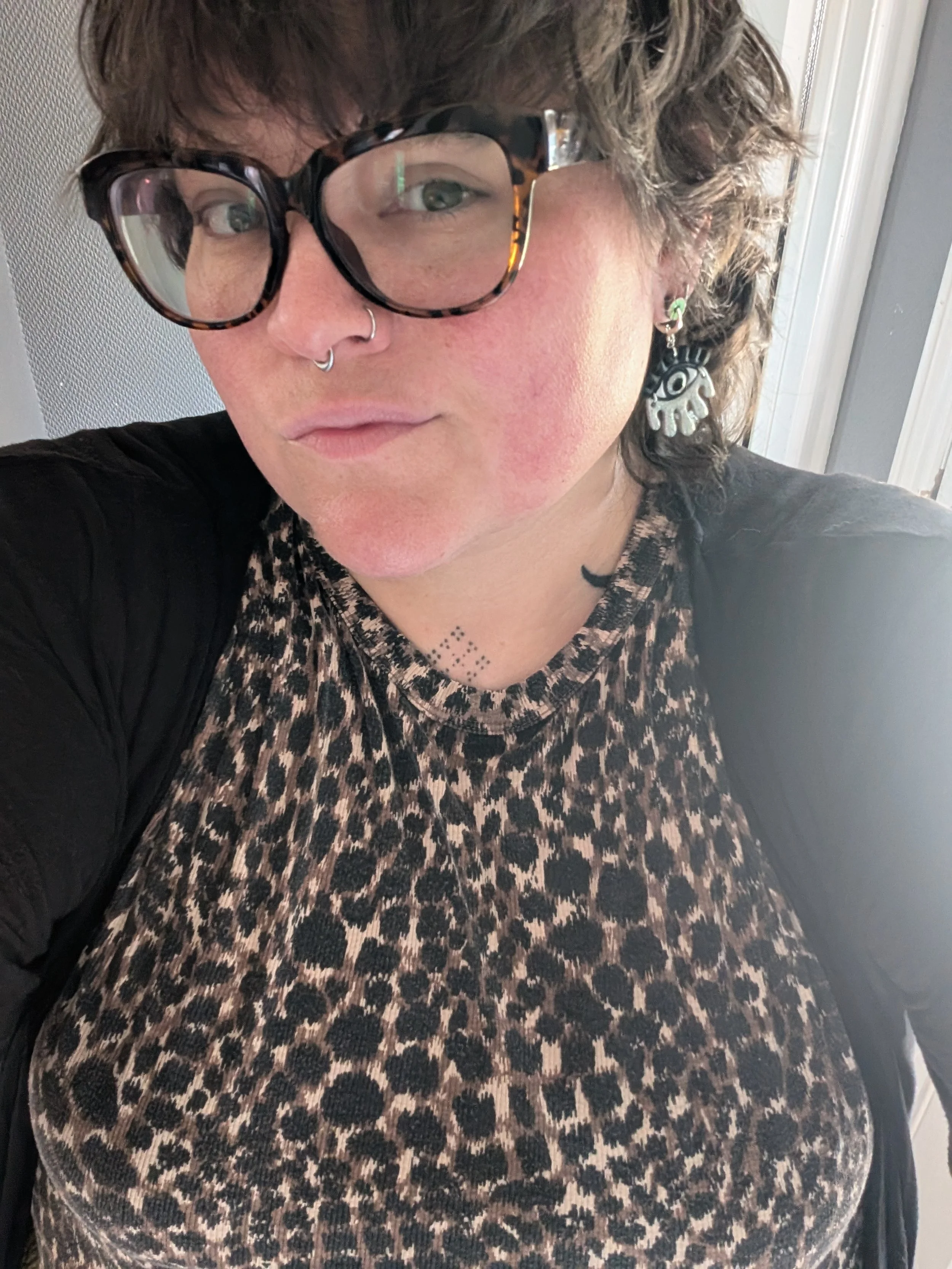 A woman with curly brown hair and glasses with tortoise shell frames, wearing a leopard print top, earrings, and a nose ring, looks at the camera.
