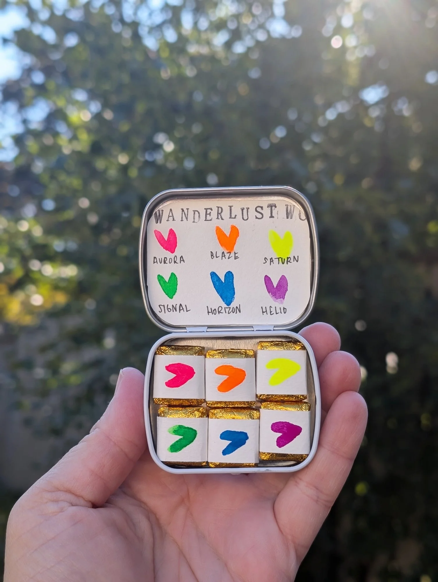 A hand holding a small rectangular box with colorful hearts and words like Aurora, Blaze, Saturn, Signal, Horizon, and Helio written inside the box, with a blurred outdoor background of trees and sunlight.