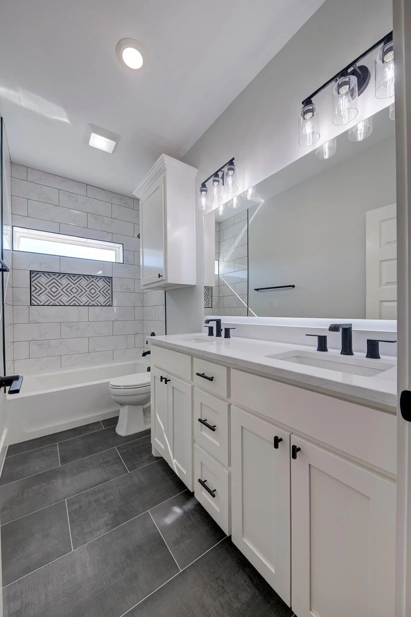 A modern bathroom with a white double vanity, black fixtures, and a large mirror. The shower area has white tiles with gray veining, a small horizontal window, and a decorative niche. The floor is dark gray tile.