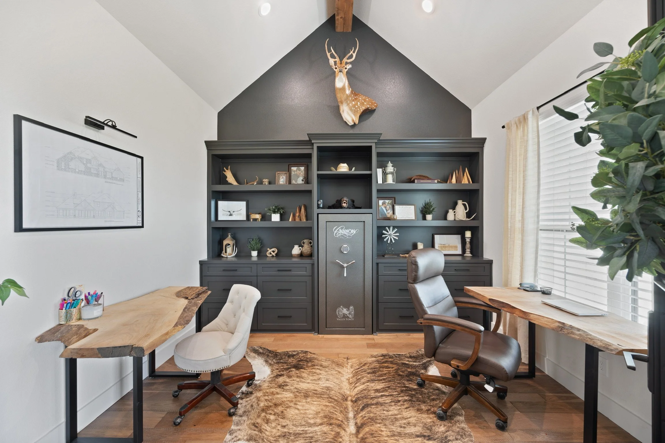 A modern home office with a high vaulted ceiling featuring a mounted deer head and dark gray shelving unit. The room has wooden surfaces, a cowhide rug, and large windows with blinds, complemented by green plants.