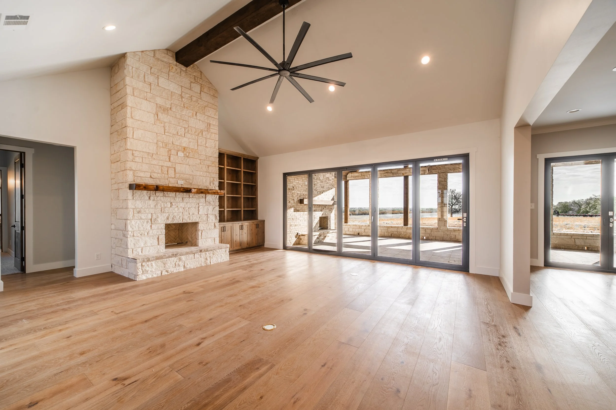 Bright living room with wooden floors, large glass sliding doors to a patio, a stone fireplace with a wooden mantle, and a ceiling fan with multiple blades.