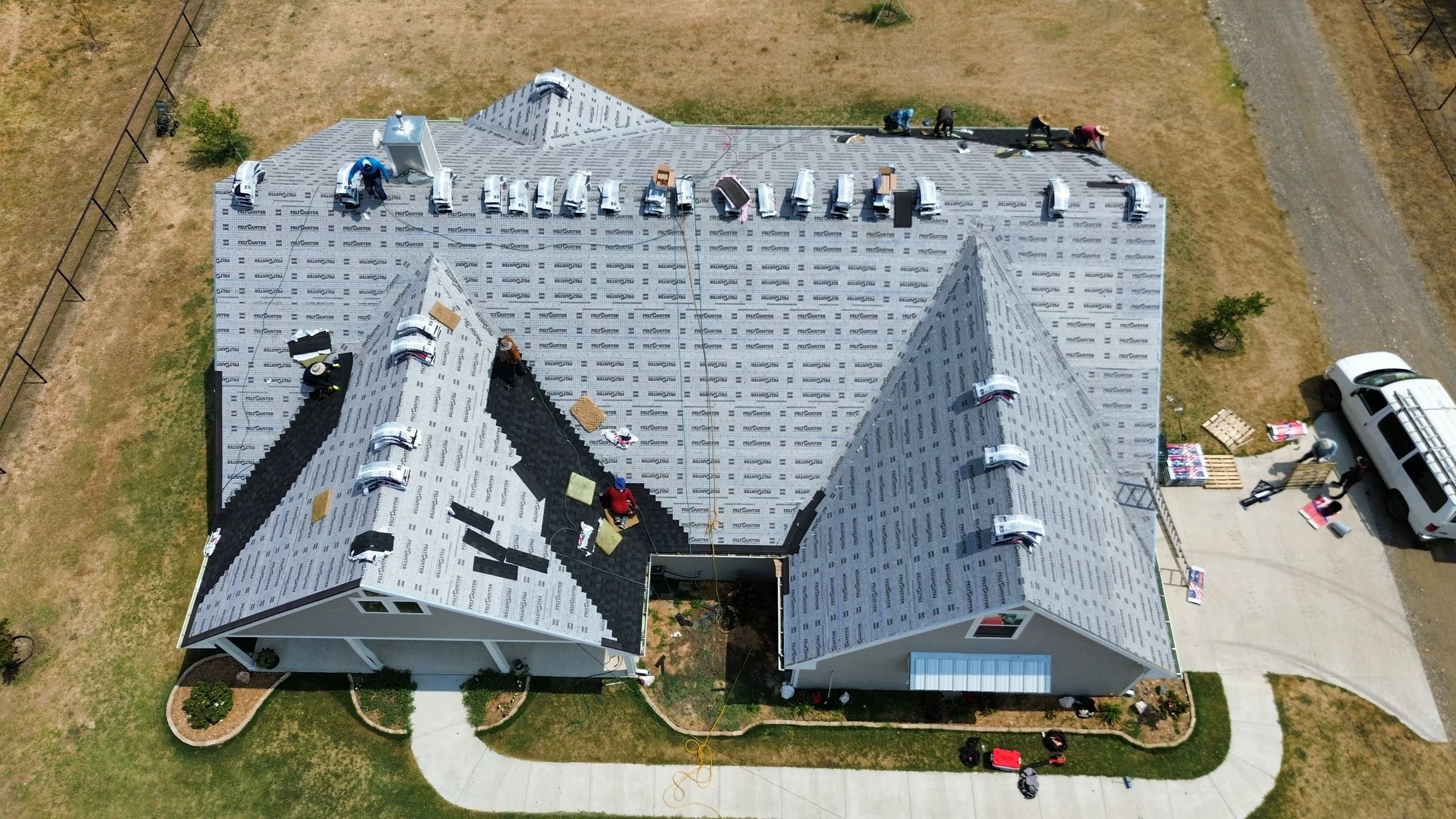 Aerial view of a house under construction, with workers installing roofing shingles on the roof, which is covered with FelteCuster roofing material.