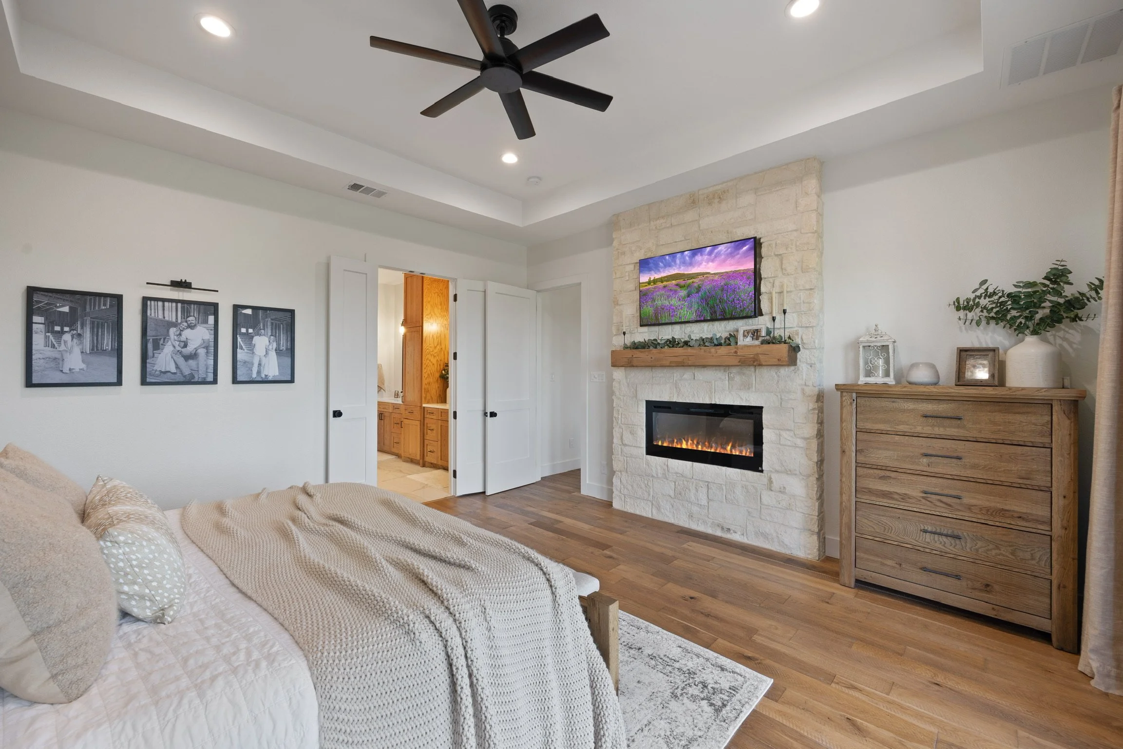 Living room with a beige couch, wood furniture, a stone fireplace with a mounted TV displaying a lavender field scene, and wall decorations including black-and-white family photos.