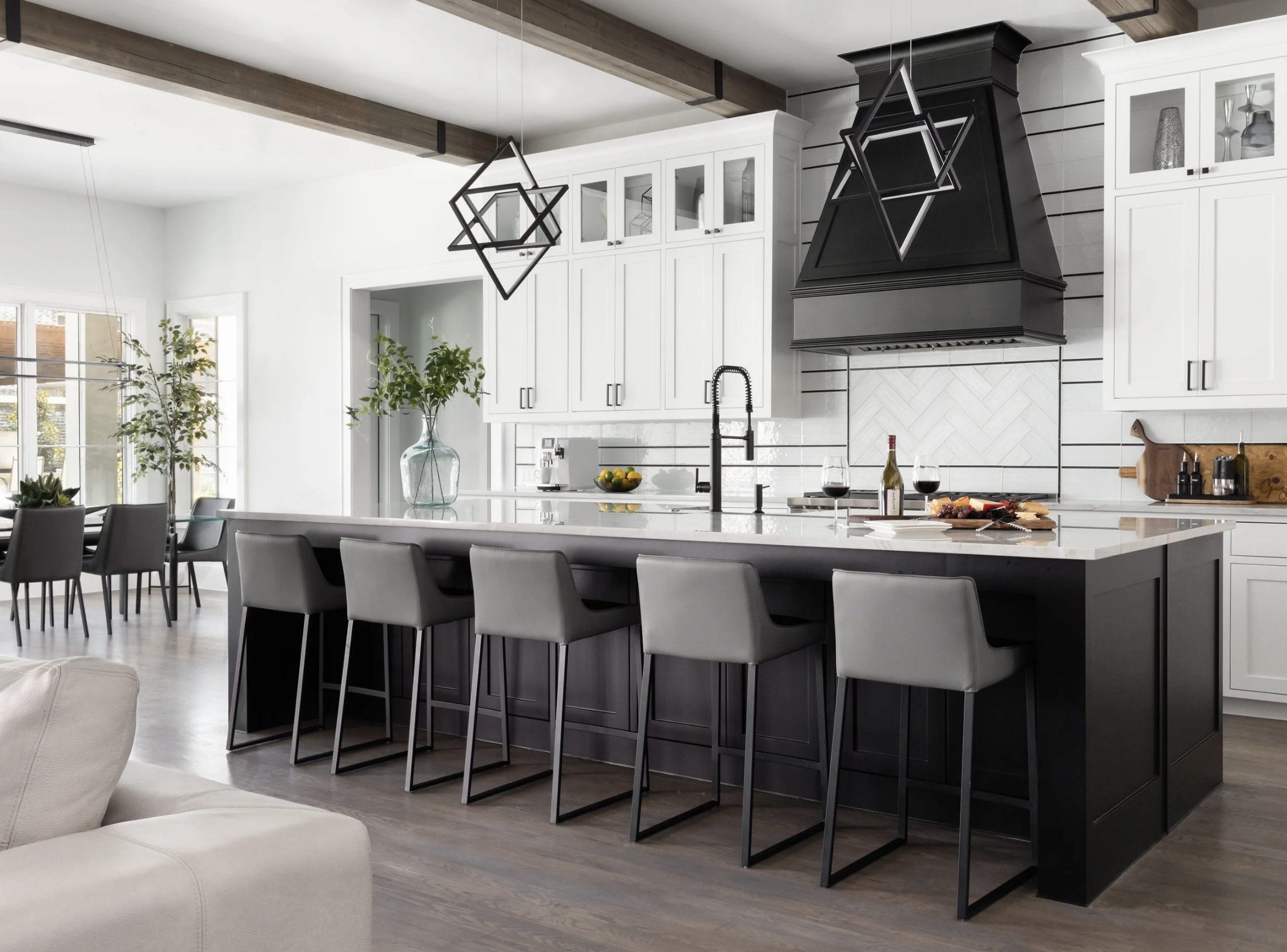 Modern kitchen with white cabinets, black range hood, island with barstools, black geometric pendant lights, glassware, wine bottles, and a vase with greenery.