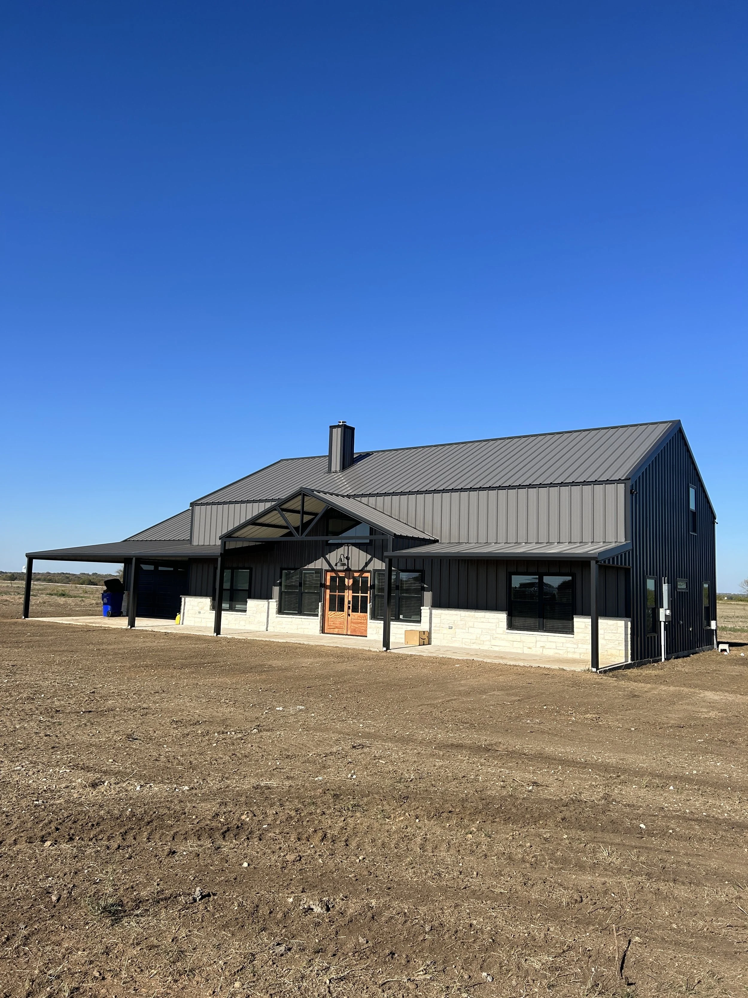 A modern black house with a gray metal roof in a wide open rural area under a clear blue sky.