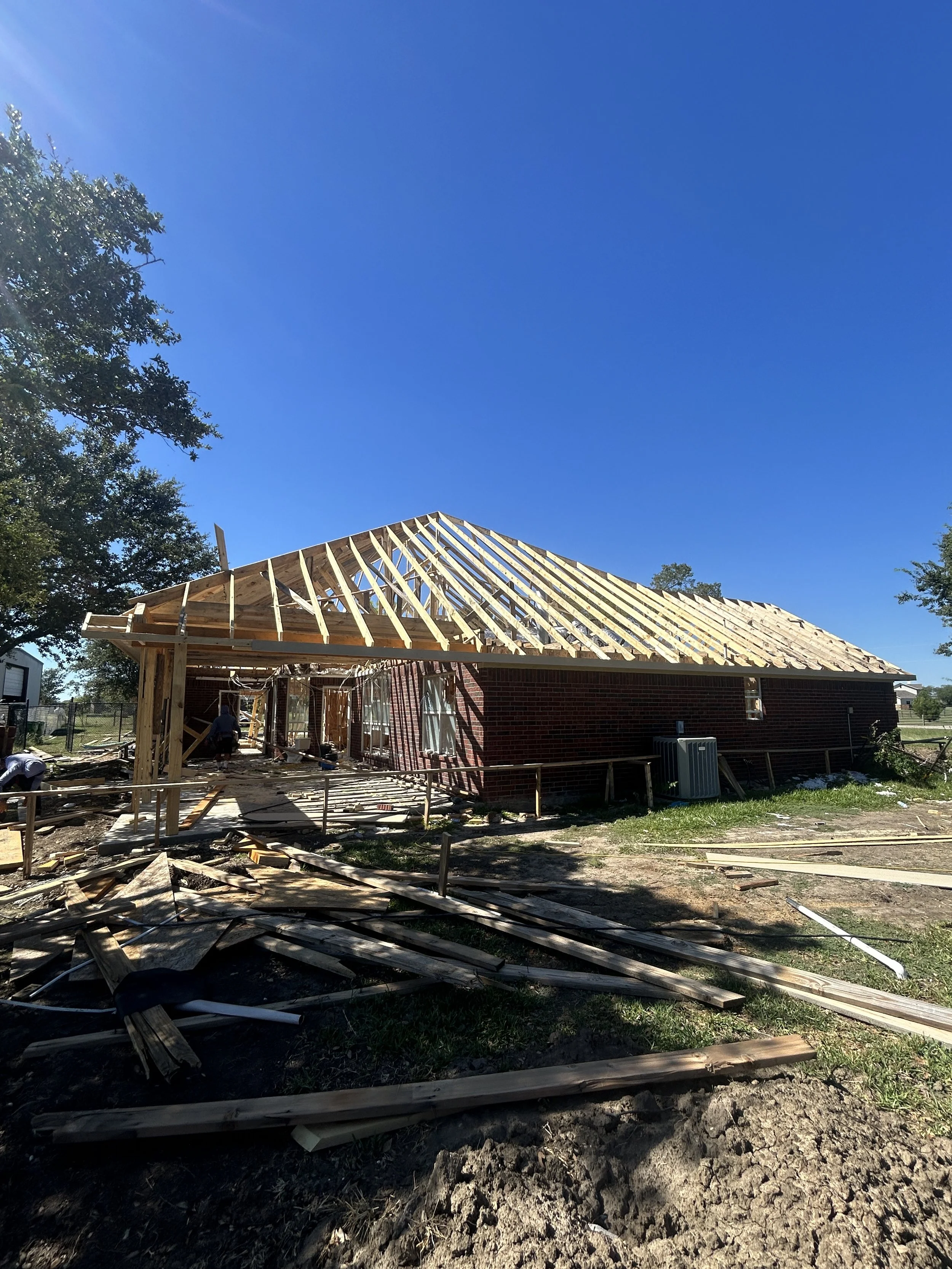 Construction site of a house with a wooden roof frame, brick walls, and construction workers working on-site, surrounded by scattered wooden planks and tools, under a clear blue sky with trees in the background.