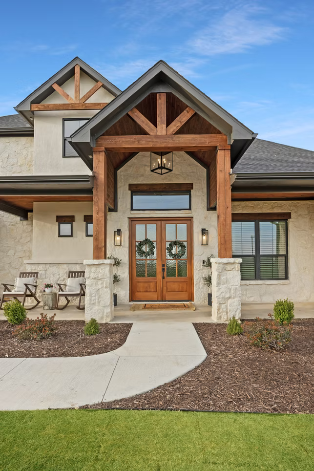 Front entrance of a modern house with wooden beams, double wooden doors decorated with holiday wreaths, stone pillars, and a porch with outdoor chairs and plants.