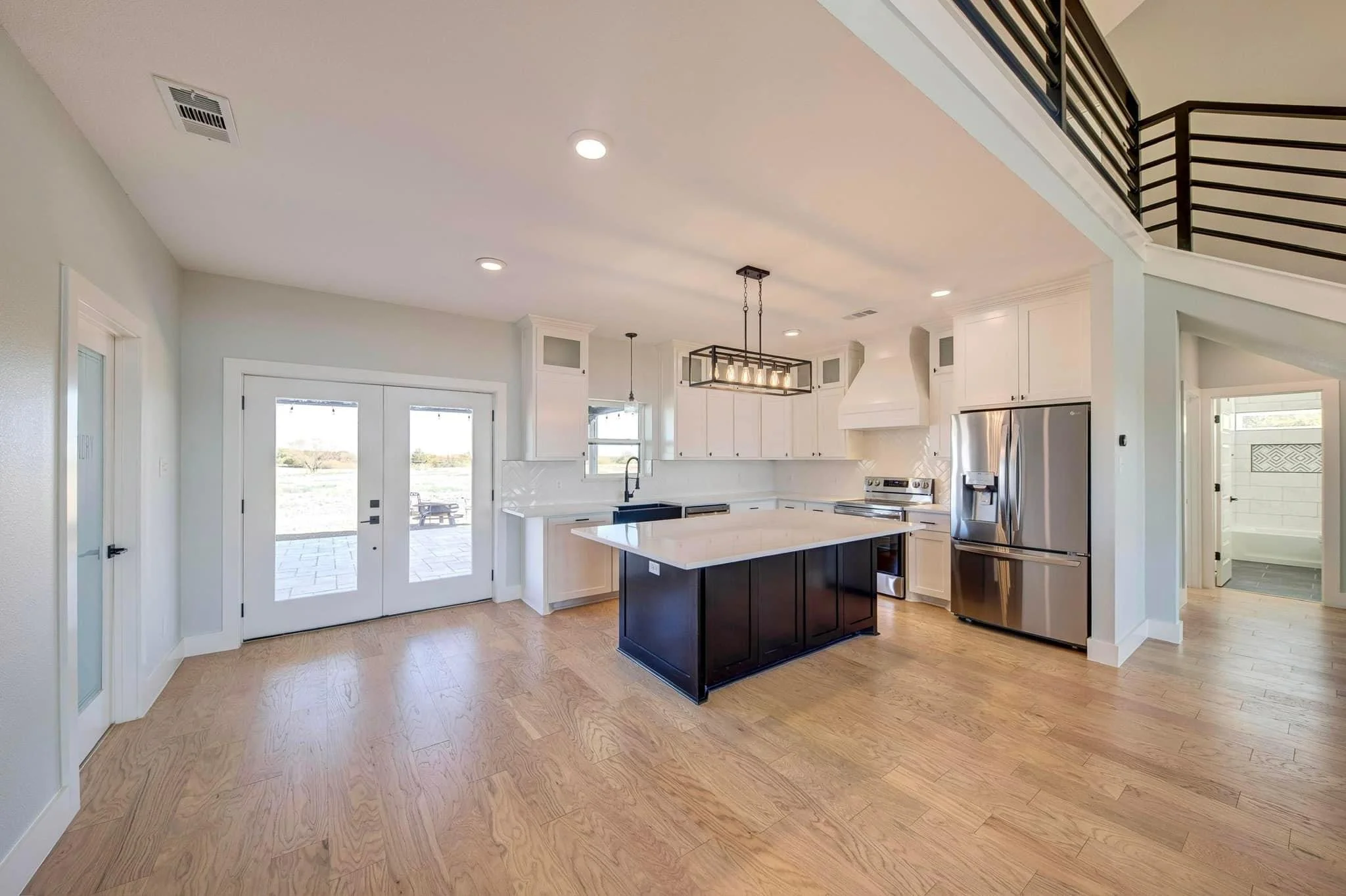 Interior view of a modern kitchen with white cabinets, a kitchen island with a dark base, a stainless steel refrigerator, and large windows for natural light.