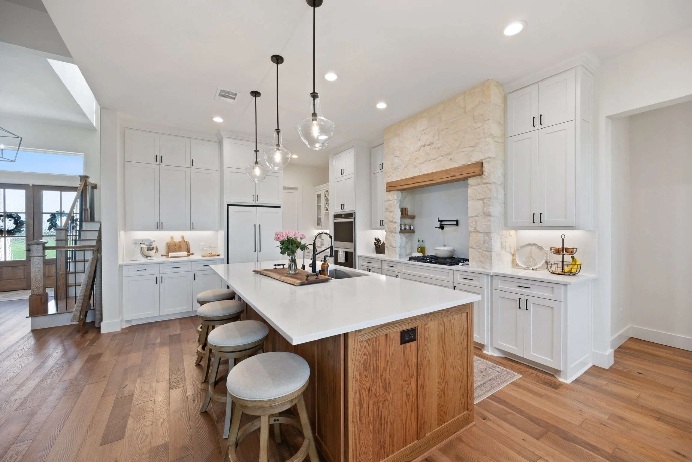 Modern kitchen with white cabinets, a large island with a white countertop, wooden accents, hanging pendant lights, and a stone fireplace overhead.