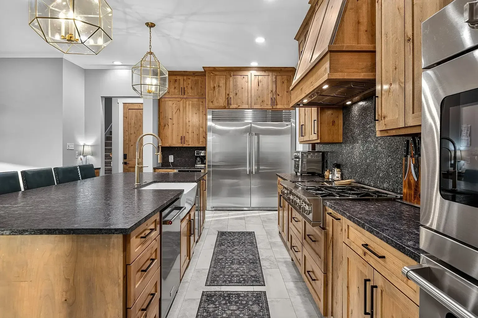 Modern kitchen with wooden cabinets, black countertops, stainless steel appliances including a large refrigerator and stove, and decorative pendant lights over a dark island with a sink.