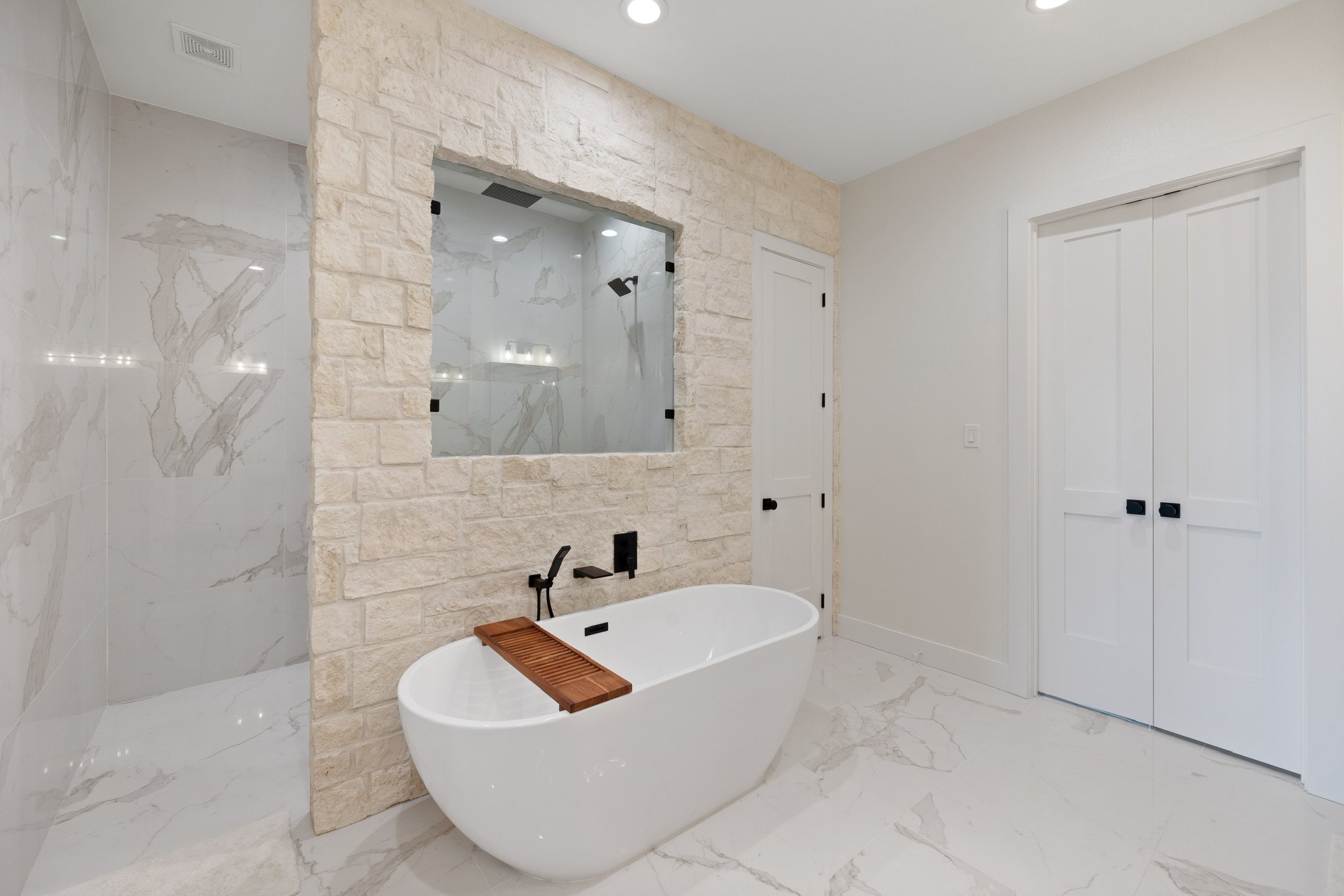 Modern bathroom with a white free-standing bathtub, a stone accent wall with a window, marble tiles, a wooden bath tray, and a white double-door closet.