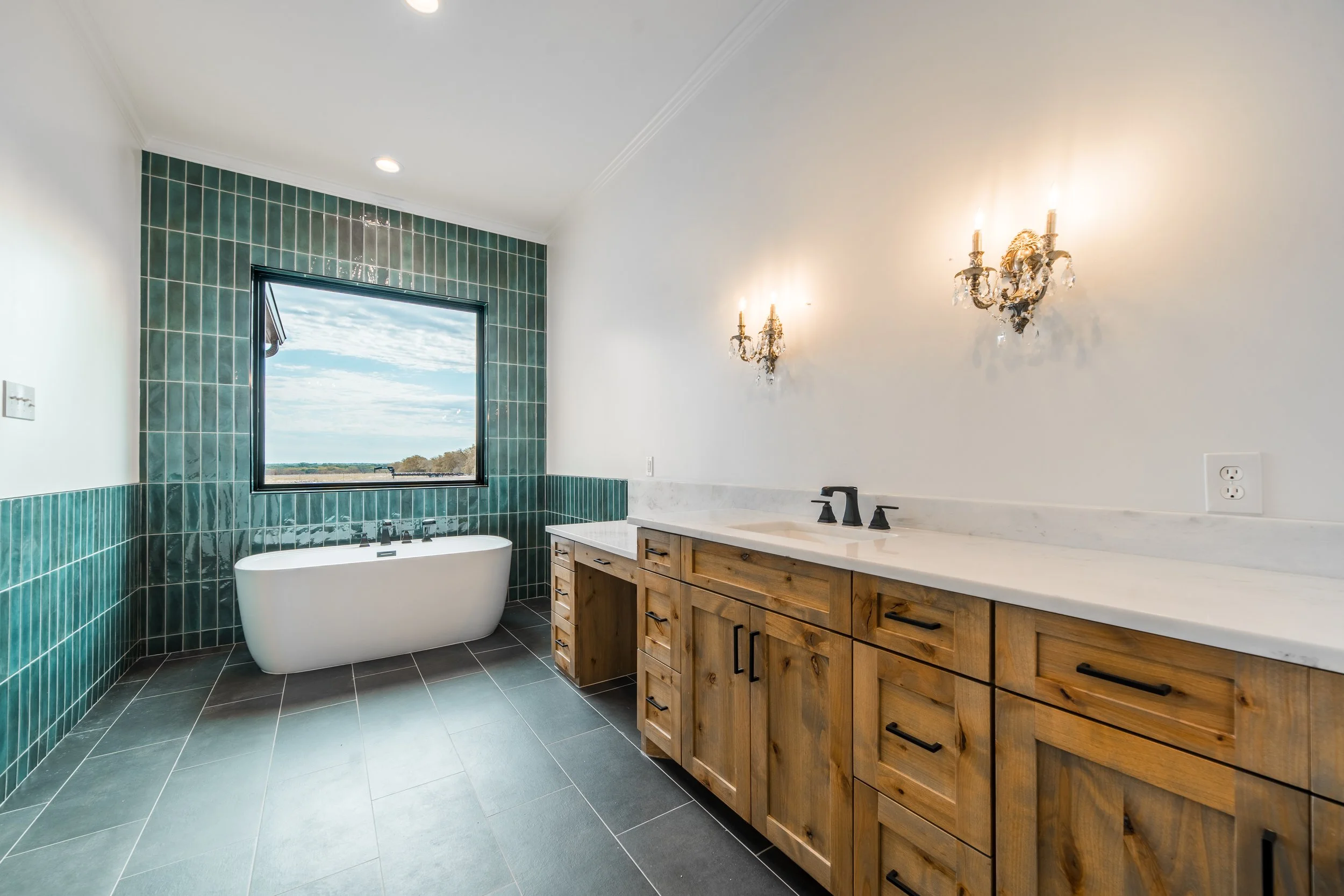 Modern bathroom with green tiled walls, a freestanding bathtub, a wooden vanity with a marble countertop, a black faucet, and decorative wall sconces, with a window showing an outdoor landscape.