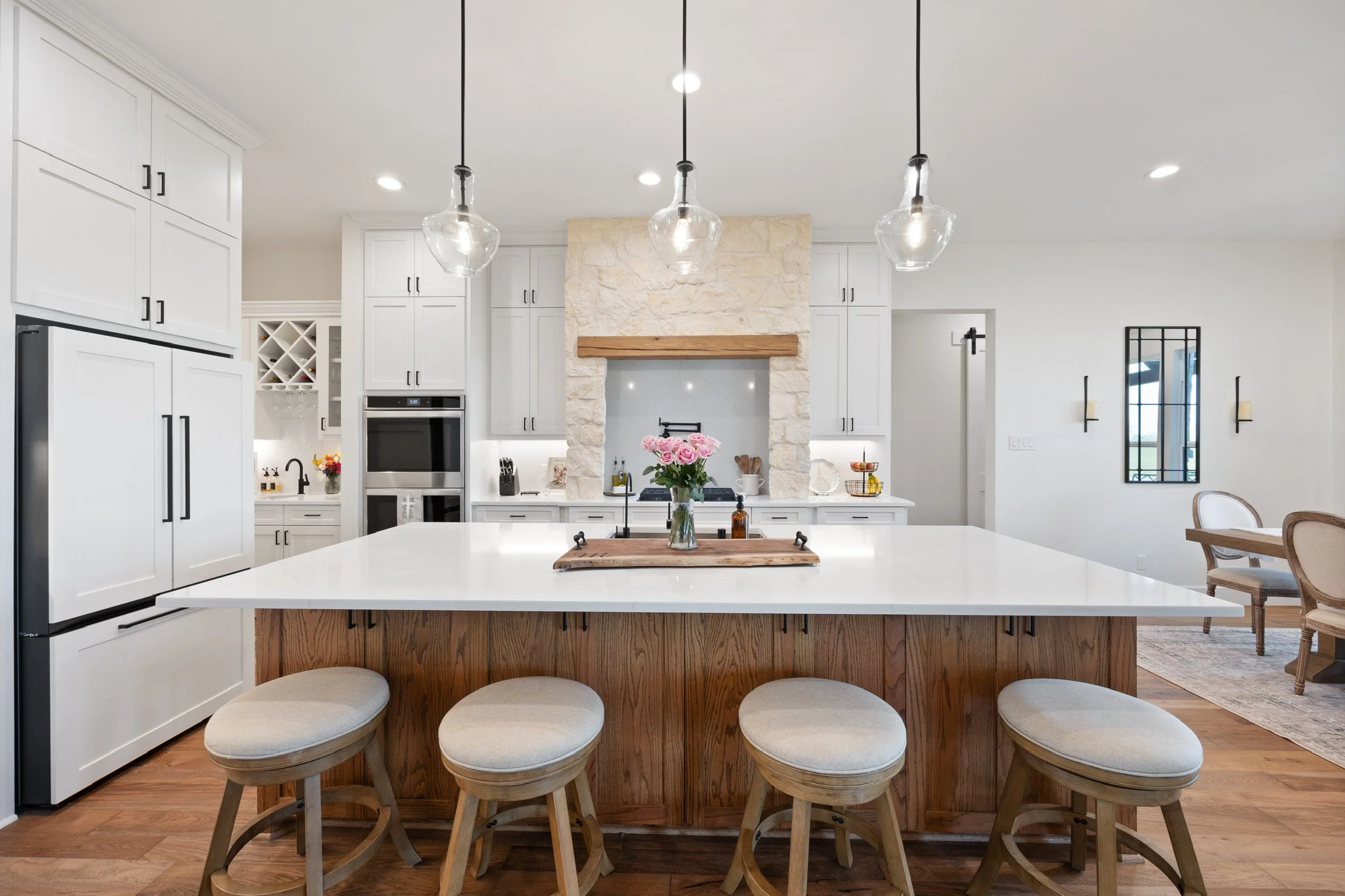 Modern kitchen with white cabinets, a stone fireplace, a large central island with a vase of pink flowers, pendant lights, and a dining area visible in the background.