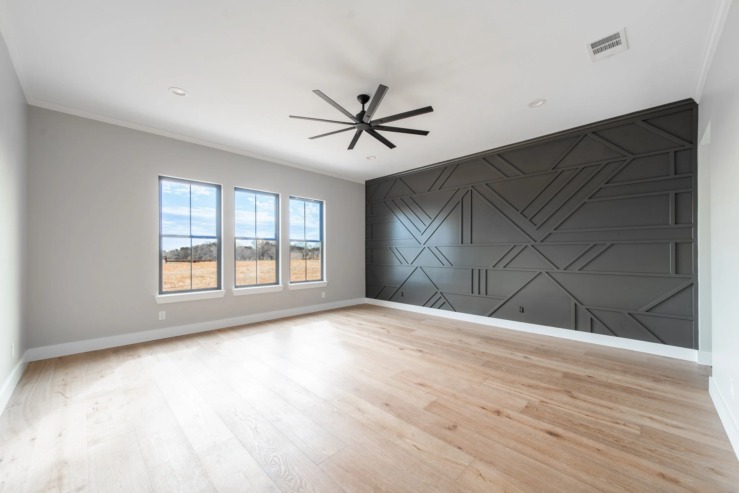 Empty room with three windows, light wood flooring, gray painted wall with geometric patterns, white ceiling, and a ceiling fan.