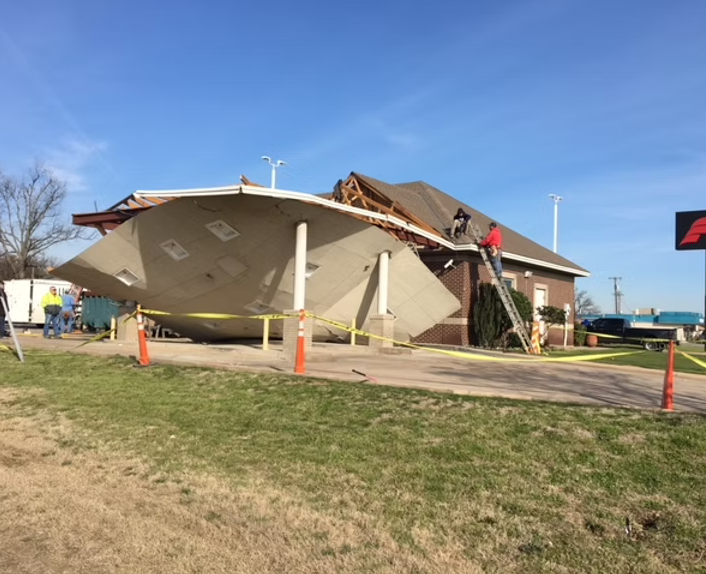 A building with a partially collapsed roof and a large part of the front wall knocked over, surrounded by caution tape and orange cones, with workers on site.