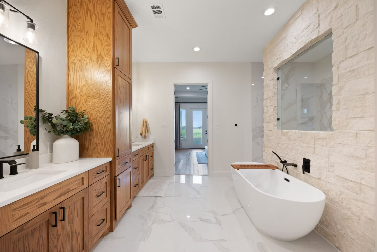 Modern bathroom with wooden cabinetry, white marble floors, a freestanding bathtub, and a stone accent wall with a window