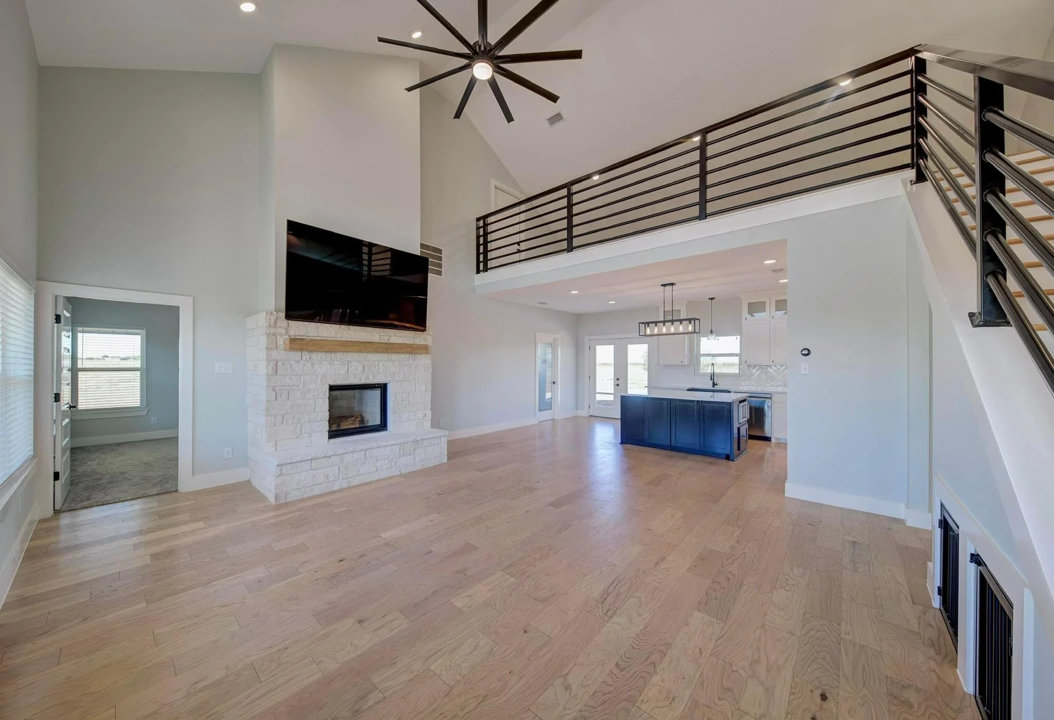 Open-concept living room with fireplace, flat-screen TV, high ceiling, and modern lighting. Kitchen with blue island and pendant lights visible in the background.