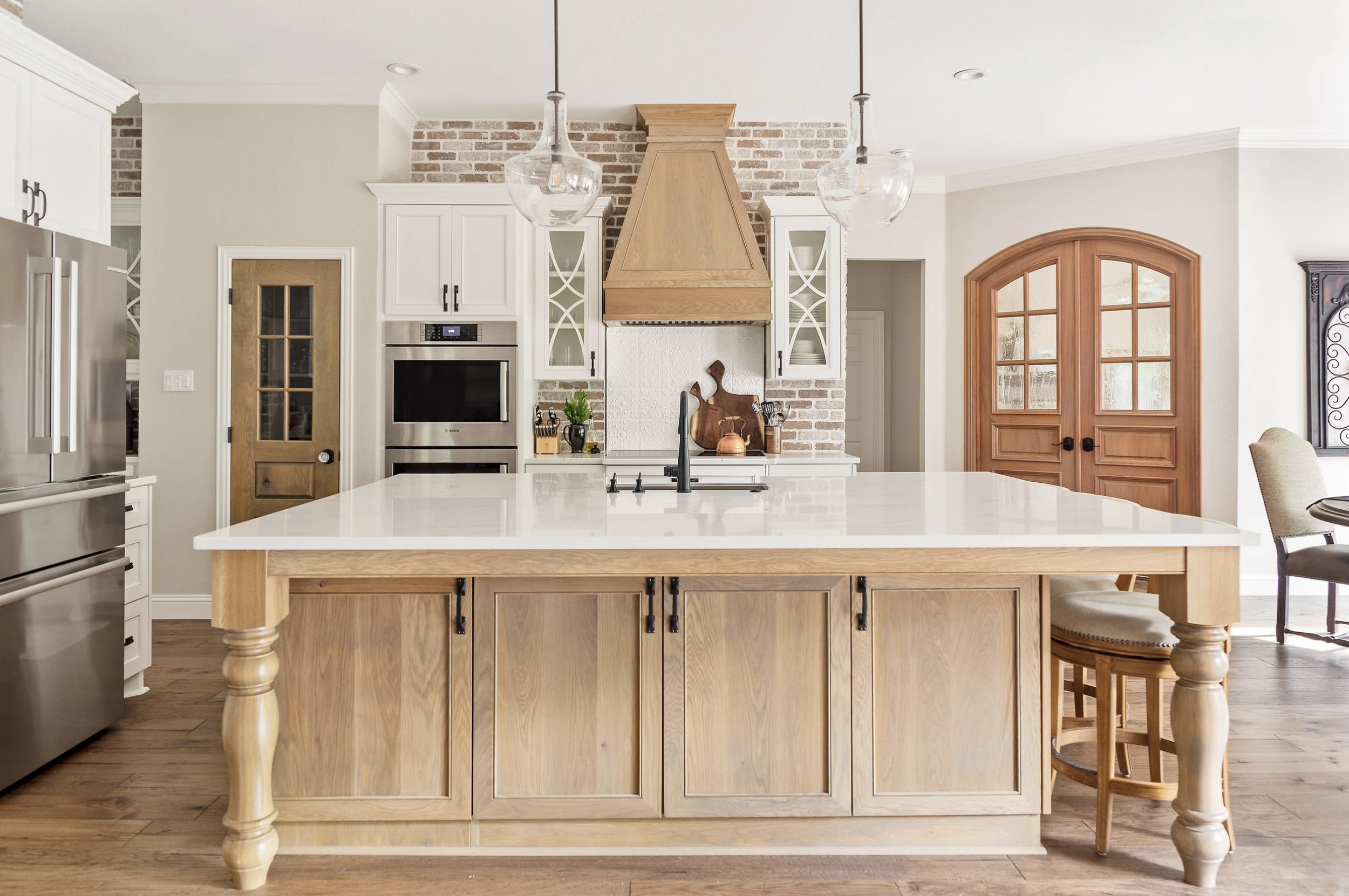 Modern kitchen with a large central island, white cabinets, stainless steel appliances, wooden accents, and a brick wall behind the stove area.