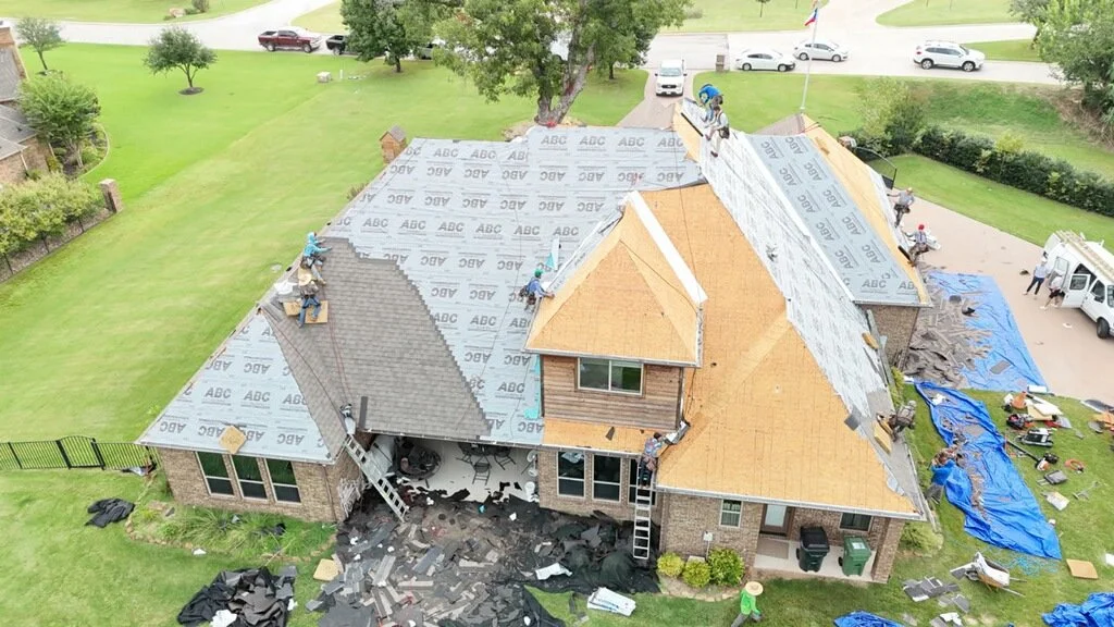 Under construction house roof with workers installing shingles, tools, and materials visible, debris on the ground, surrounded by green yard and trees.