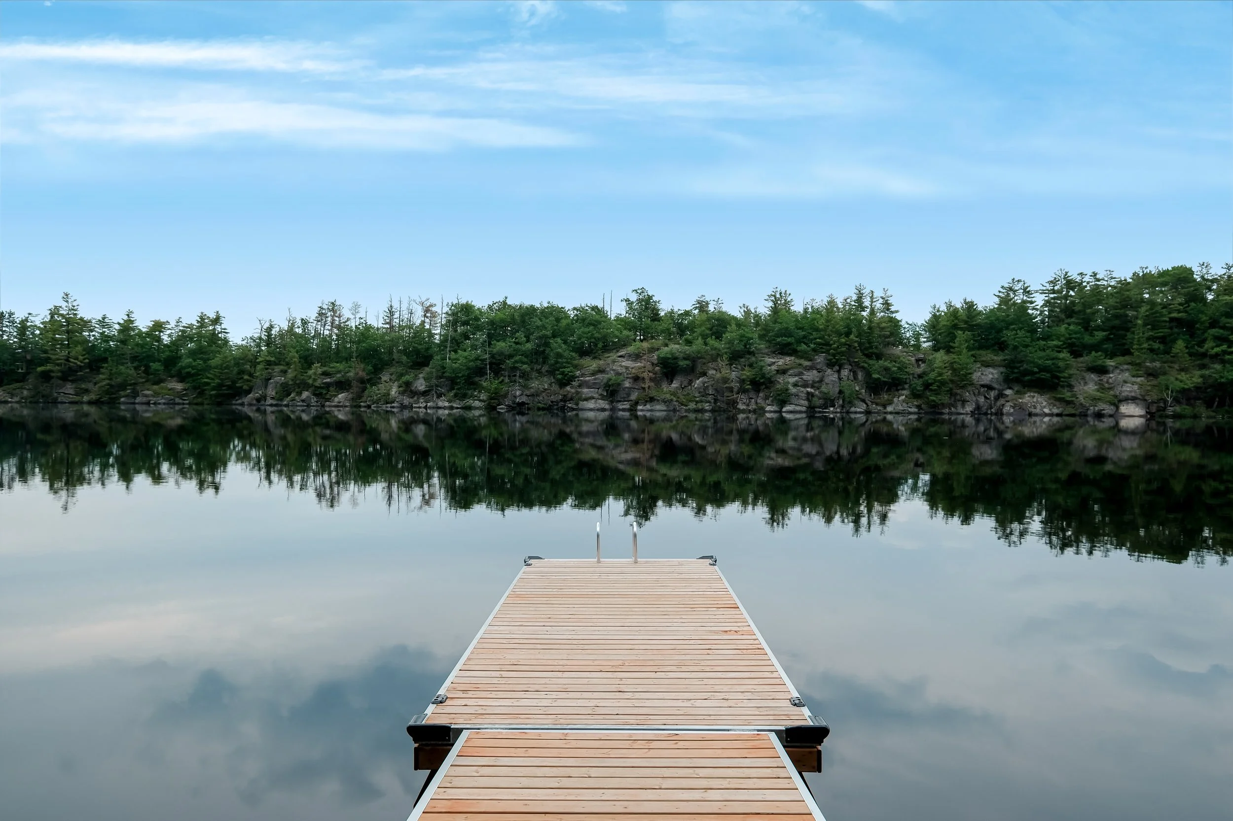 A wooden dock extends into a calm lake in Muskoka surrounded by lush green trees, with a clear blue sky reflected in the water.