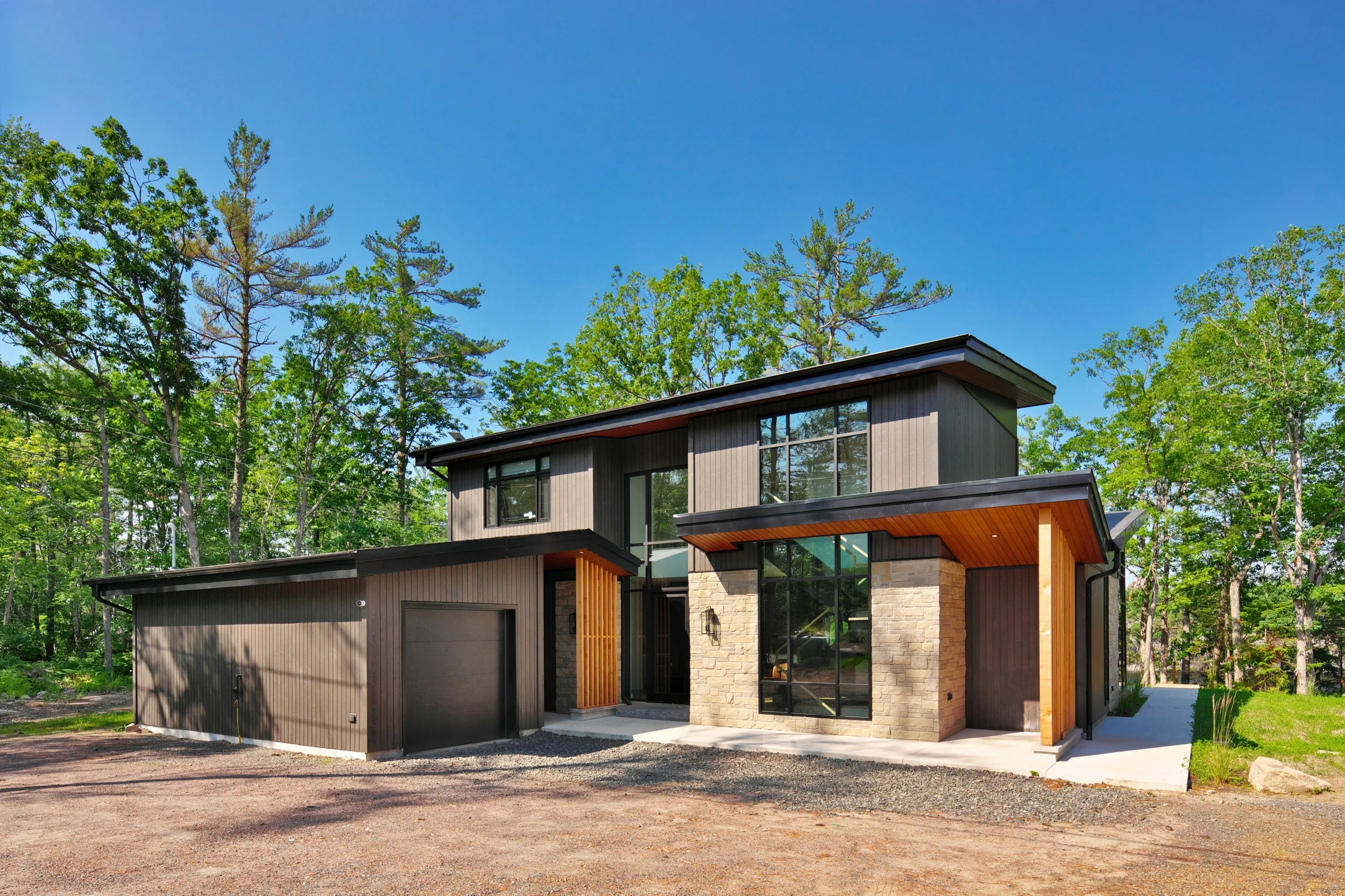 Modern Muskoka cottage with a mix of dark siding and stone exterior, large windows, and wooden accents, surrounded by green trees under a blue sky.