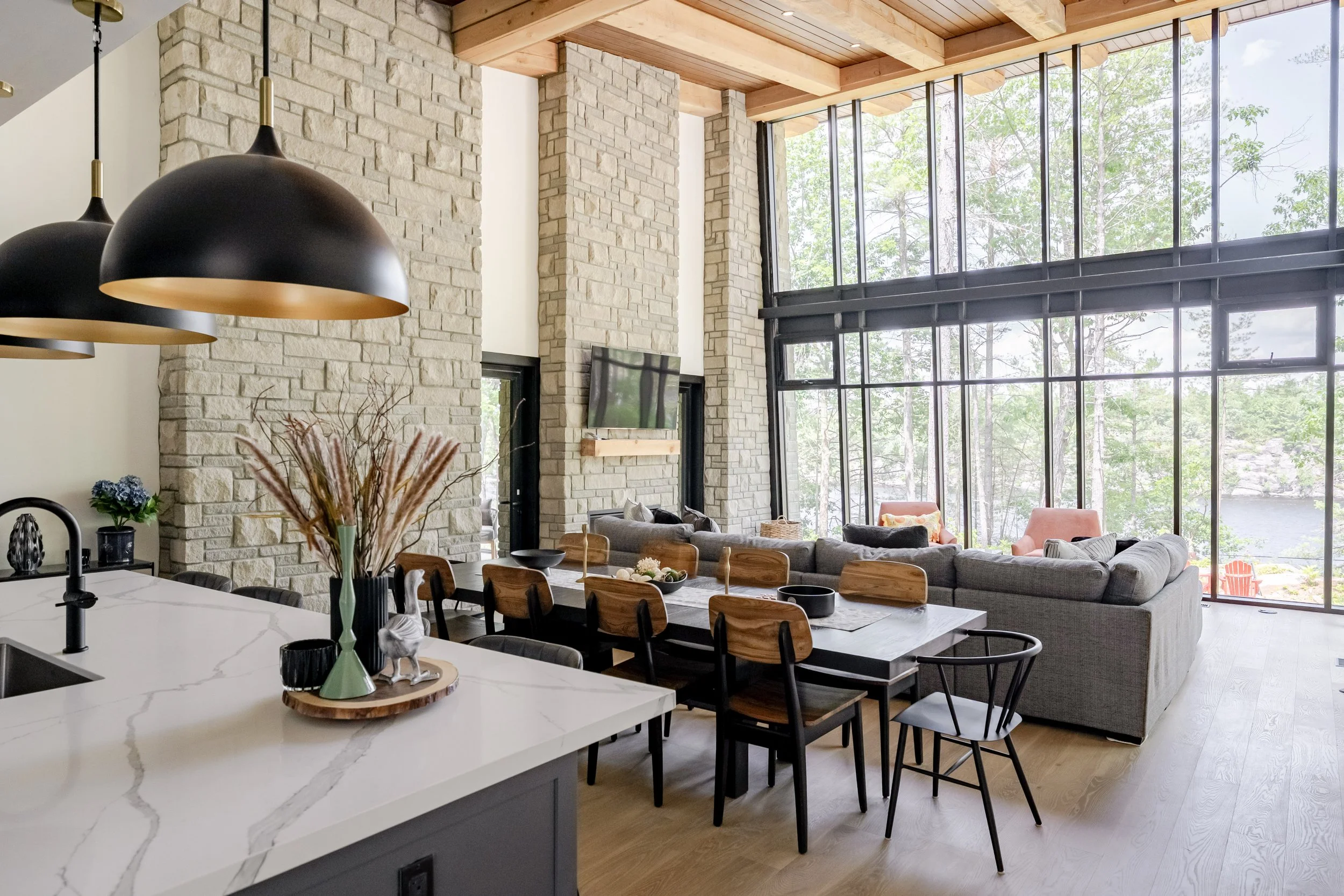 Modern living room with large floor-to-ceiling windows, stone accent walls, a gray sectional sofa, and a wooden ceiling with exposed beams.