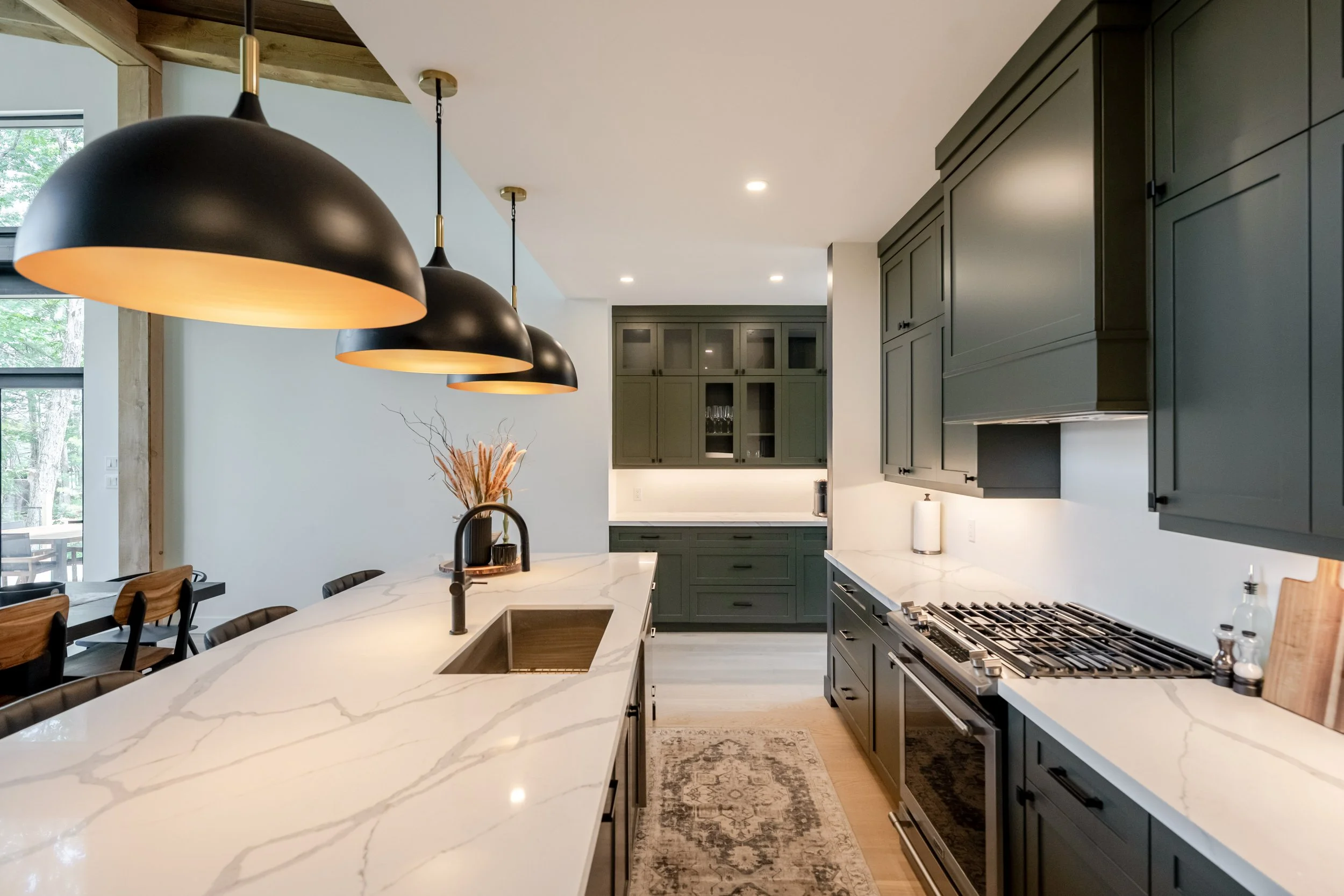 Modern kitchen with dark green cabinets, white marble island, black pendant lights, and large windows showing trees outside.