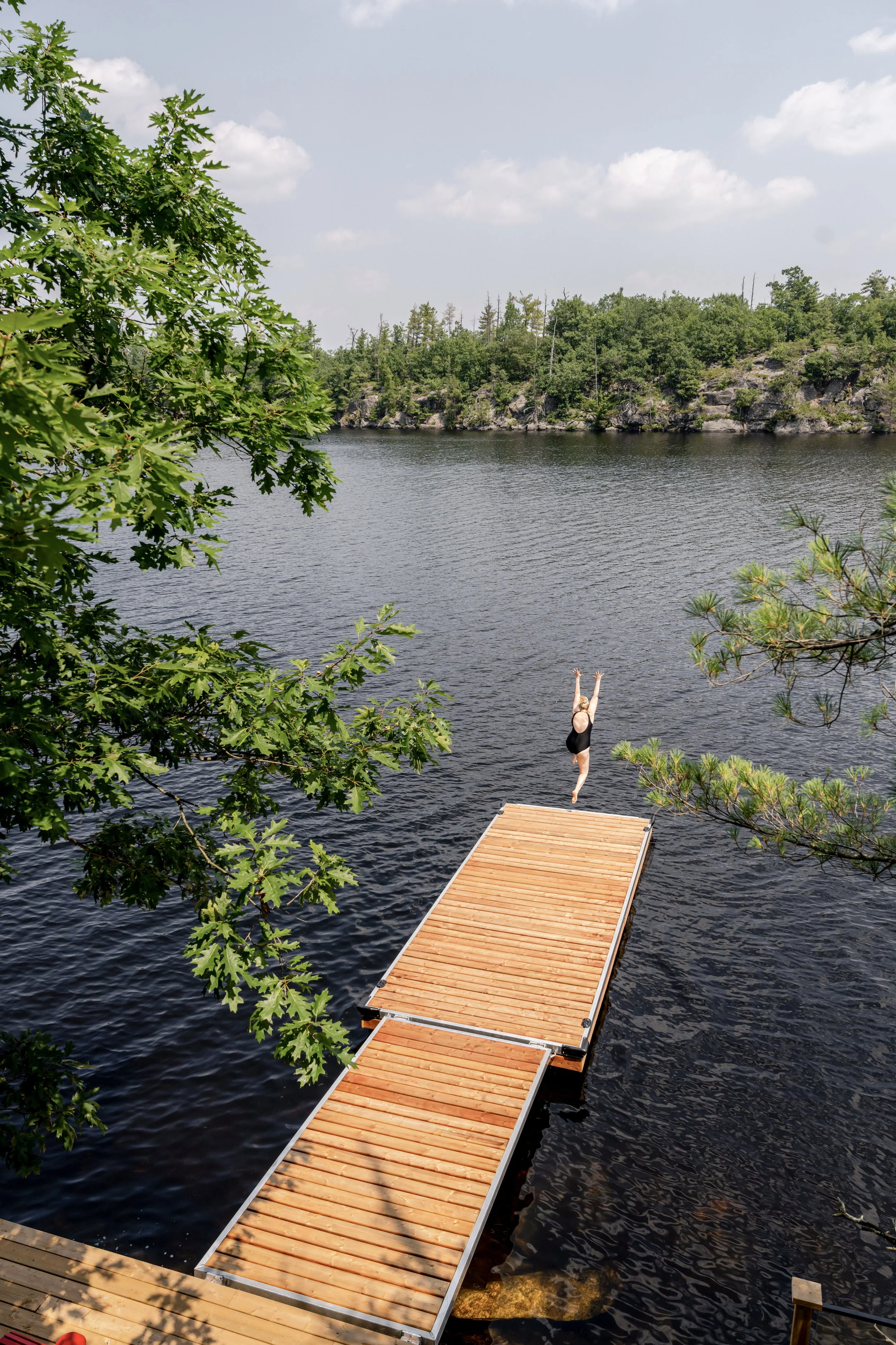 A person in a black swimsuit diving off a wooden dock into a calm lake in Gravenhurst, with trees and rocky hills in the background under a partly cloudy sky.