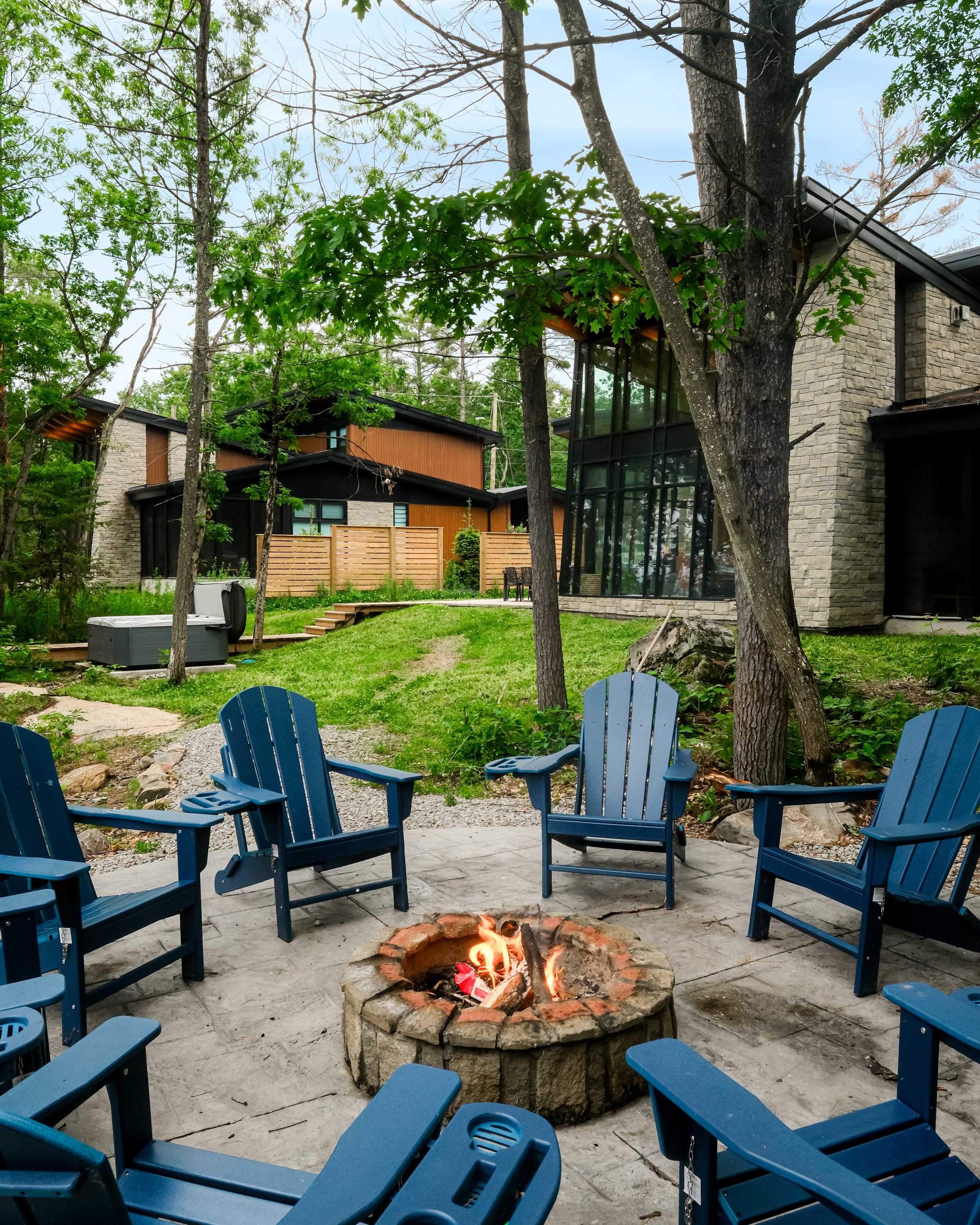 Outdoor seating area with blue Adirondack chairs arranged in a circle around a fire pit with a small fire burning. Behind, there are trees and a modern house with large windows and stone walls.