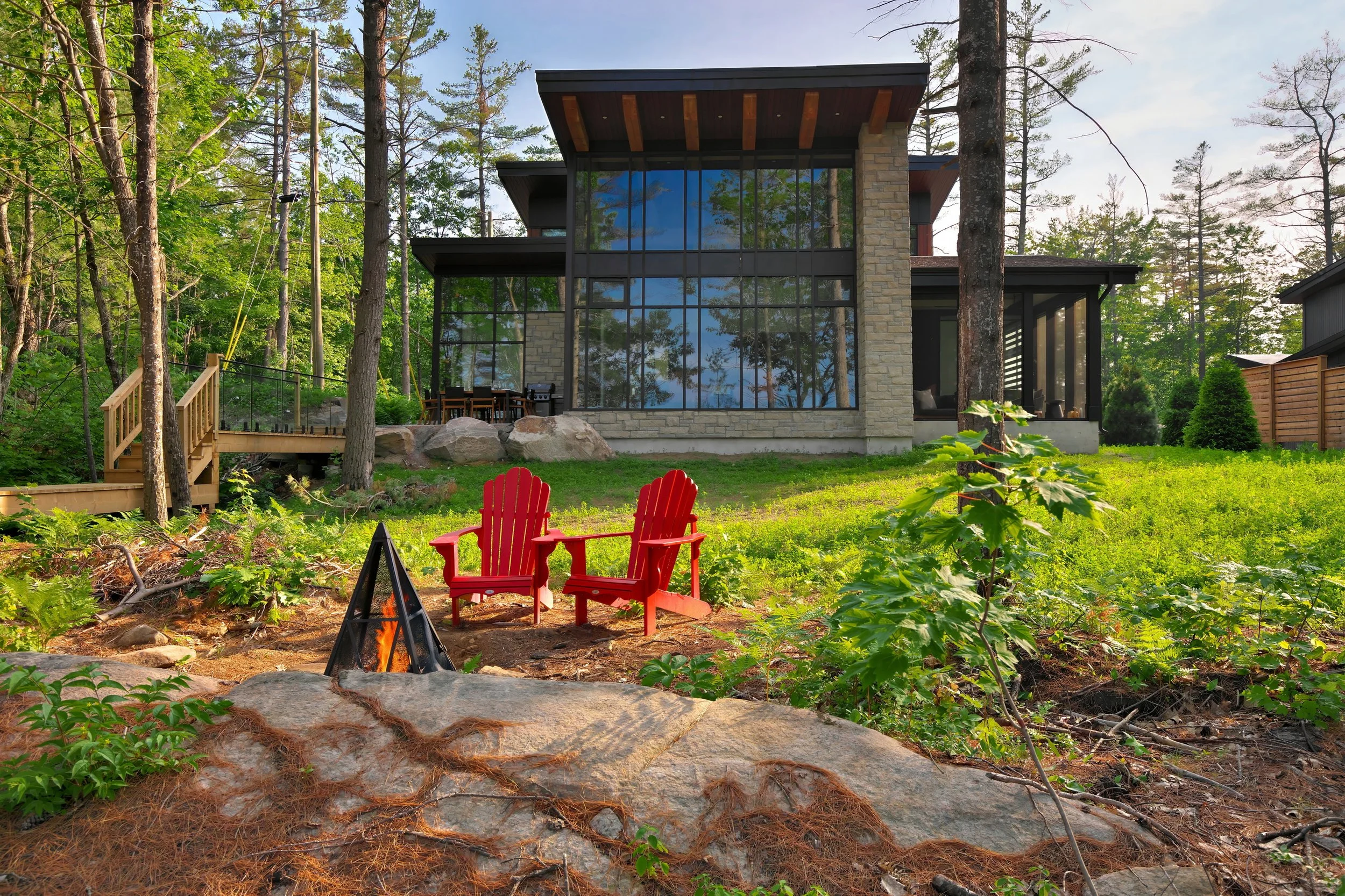 A modern house with large glass windows situated among tall trees in a wooded area. The yard features a fire pit with red Adirondack chairs, a small stone pathway, and a wooden deck with outdoor seating.