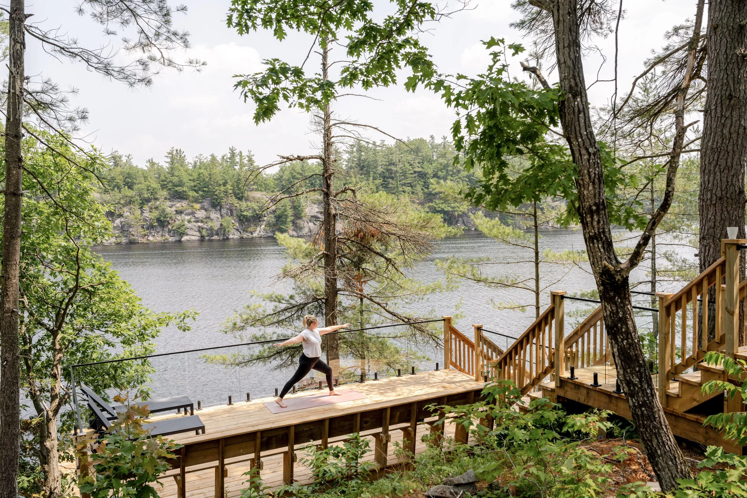 A woman practicing yoga on a wooden deck with a view of a lake, trees, and mountains, surrounded by lush green foliage. Peaceful observation deck for large gatherings or corporate retreats.