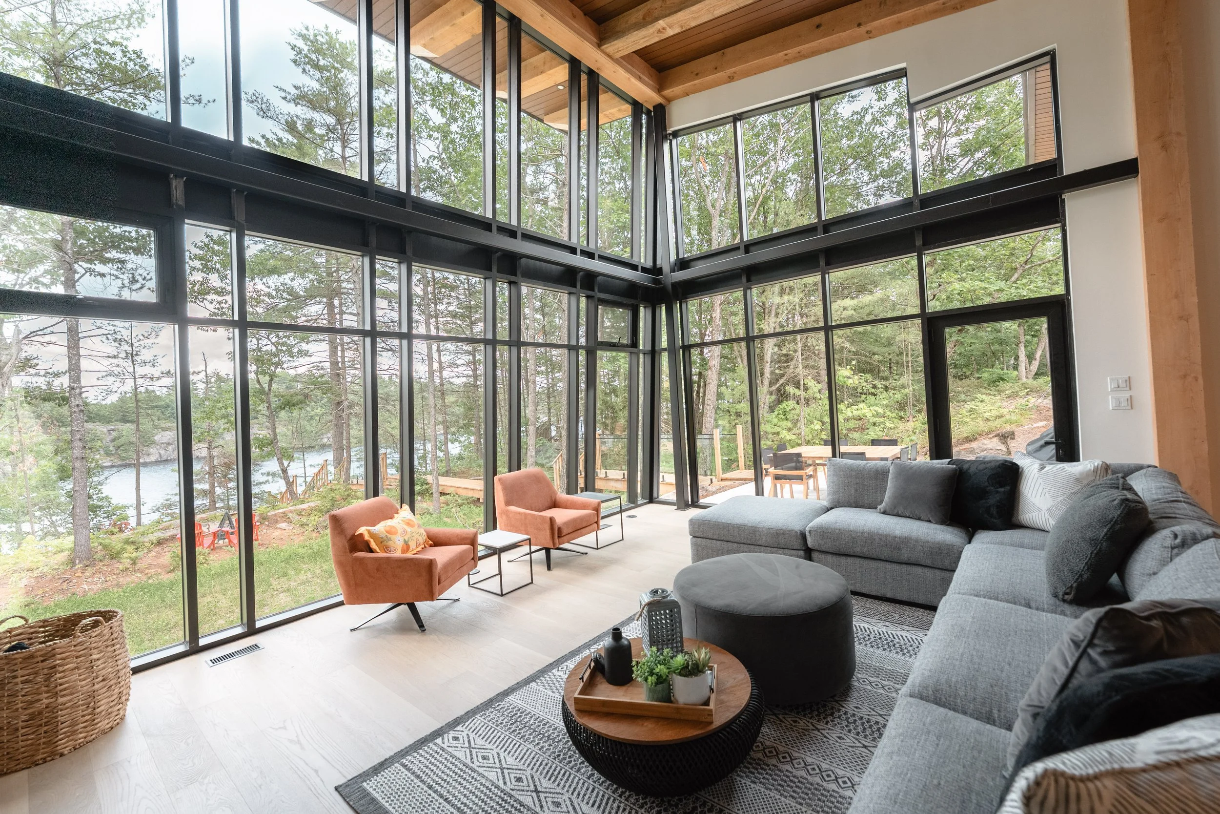 Living room with large floor-to-ceiling windows overlooking a wooded outdoor area. The room contains a gray sectional sofa, two orange armchairs with a small side table, a patterned rug, and a round black coffee table with plants. The ceiling has wooden beams.