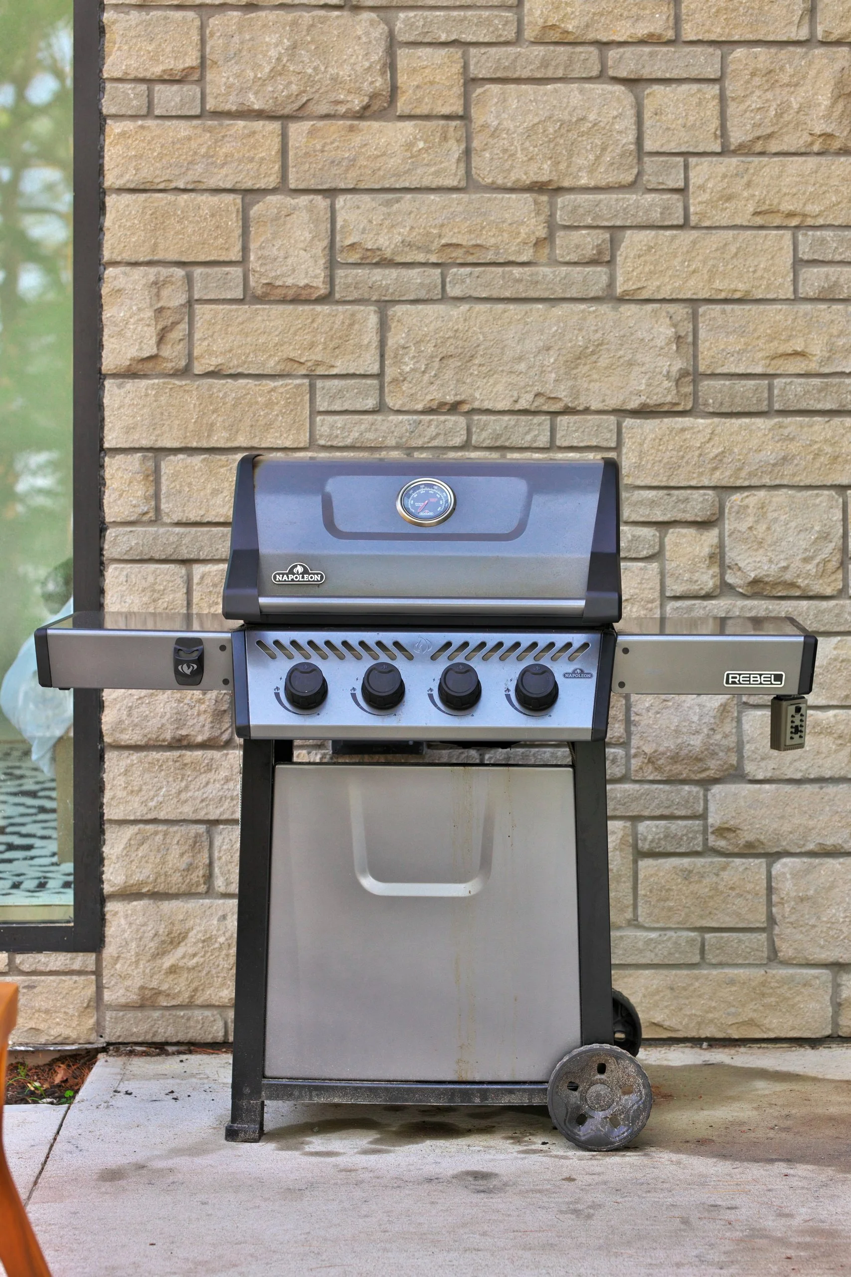A propane outdoor gas grill with four control knobs, a built-in thermometer on the lid, and wheels on the bottom, placed against a beige brick wall.
