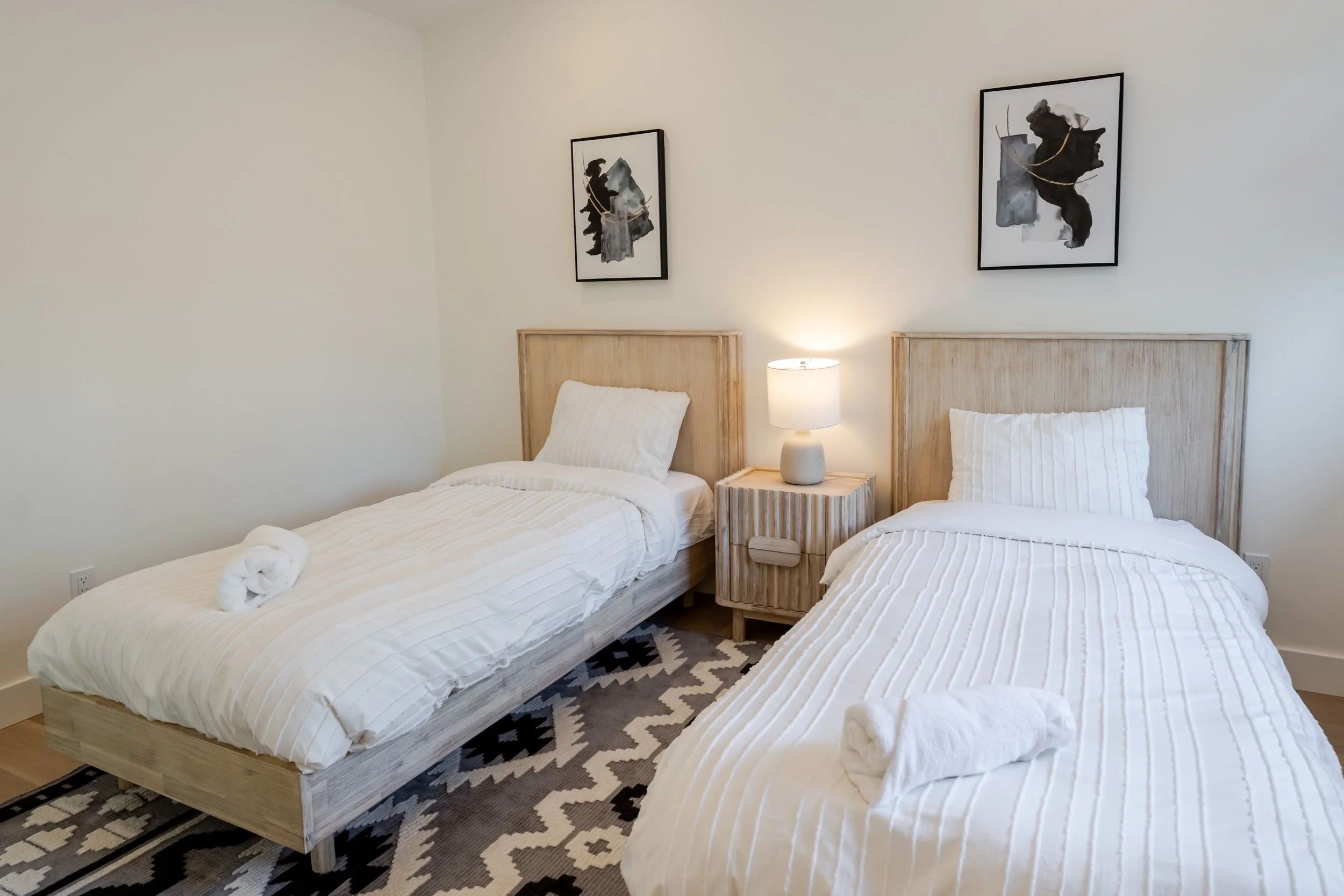 A bedroom with two twin beds featuring white striped bedding, wooden headboards, and white pillows. A small wooden nightstand with a white lamp is placed between the beds. The wall above the beds is decorated with two black and white abstract art prints, and a black and white rug with a geometric pattern lies on the floor.