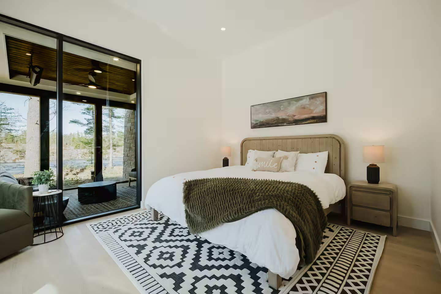 Modern bedroom with white walls, a large bed with white bedding and a dark green throw blanket, wooden headboard, two bedside tables with lamps, a painting above the bed, a patterned rug, and a glass door leading to a balcony with outdoor seating and trees outside.