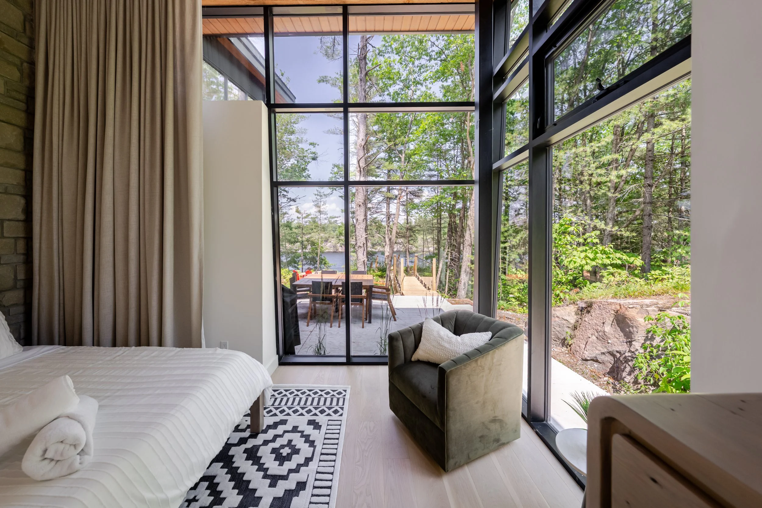 Interior of a modern room with large floor-to-ceiling windows showcasing a wooded outdoor view, a bed with white linens, a black and white geometric rug, a green armchair with a white pillow, and beige curtains.