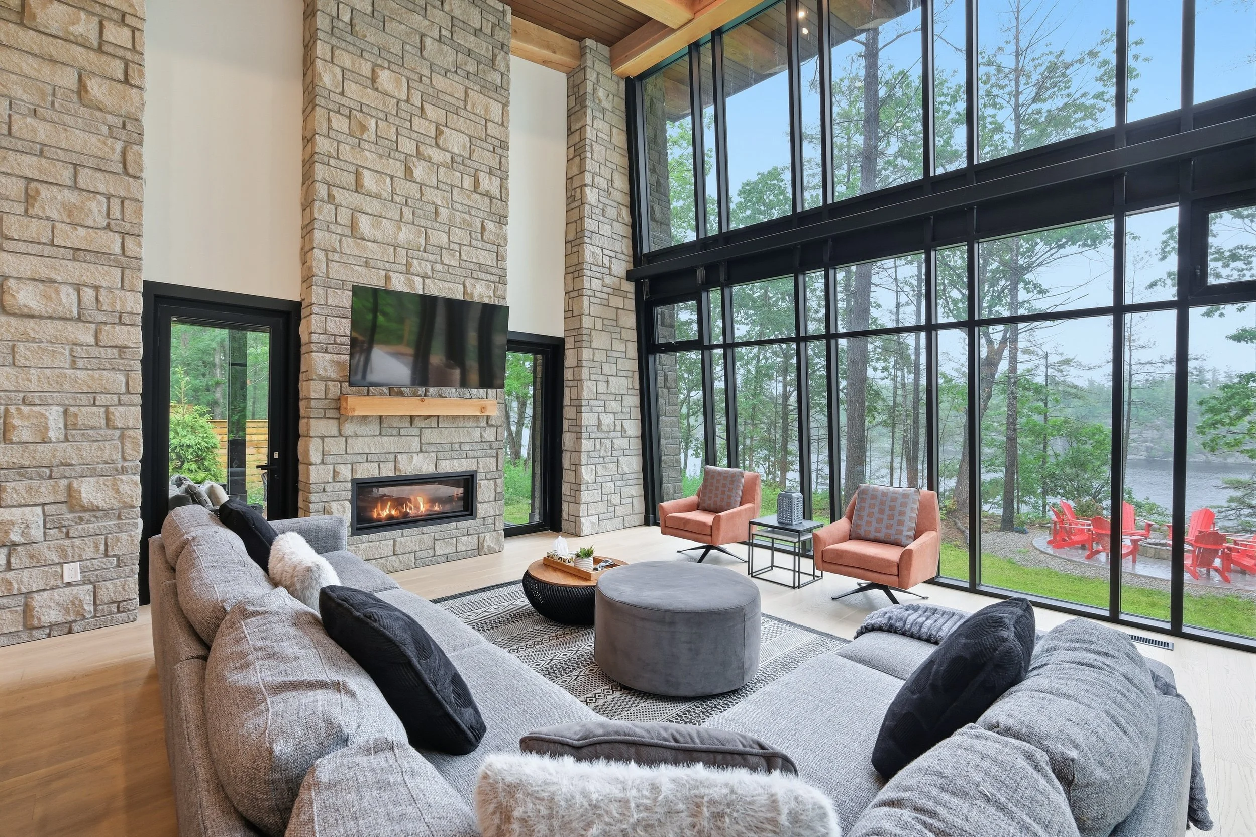 Interior of a modern living room with a high ceiling, large windows showing green trees and water outside, a stone fireplace, a wall-mounted TV, a gray sectional sofa with black and white pillows, two pink armchairs, a round gray ottoman, a small side table with a decorative item, and a view of a red outdoor patio with Adirondack chairs.