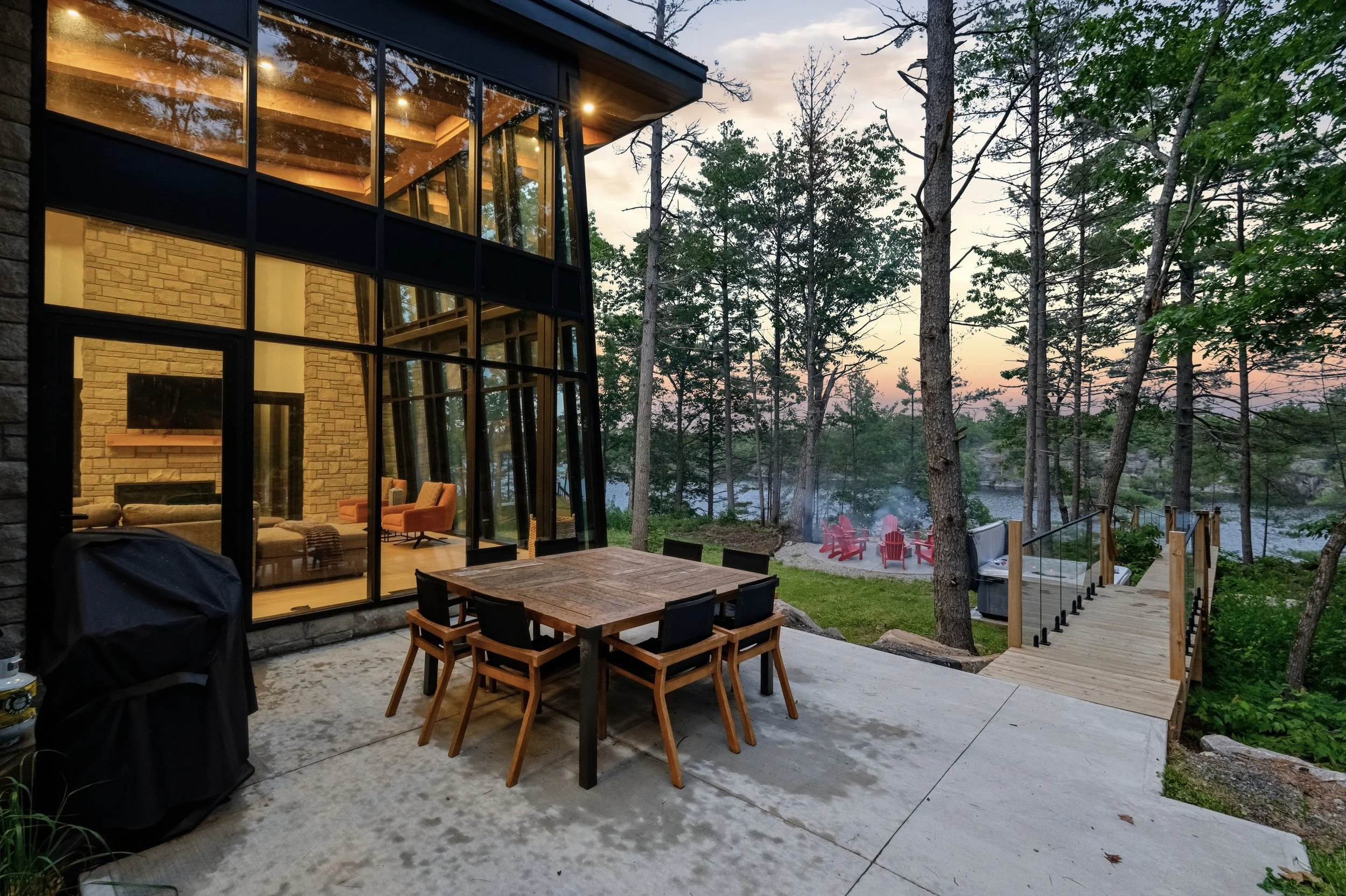A modern house with large glass windows revealing a cozy interior living room with chairs and a fireplace. In the backyard, there is a patio with a wooden dining table, a grill, and a fire pit area with red Adirondack chairs. The yard has a hillside with trees and a water body in the background, during dusk or evening.