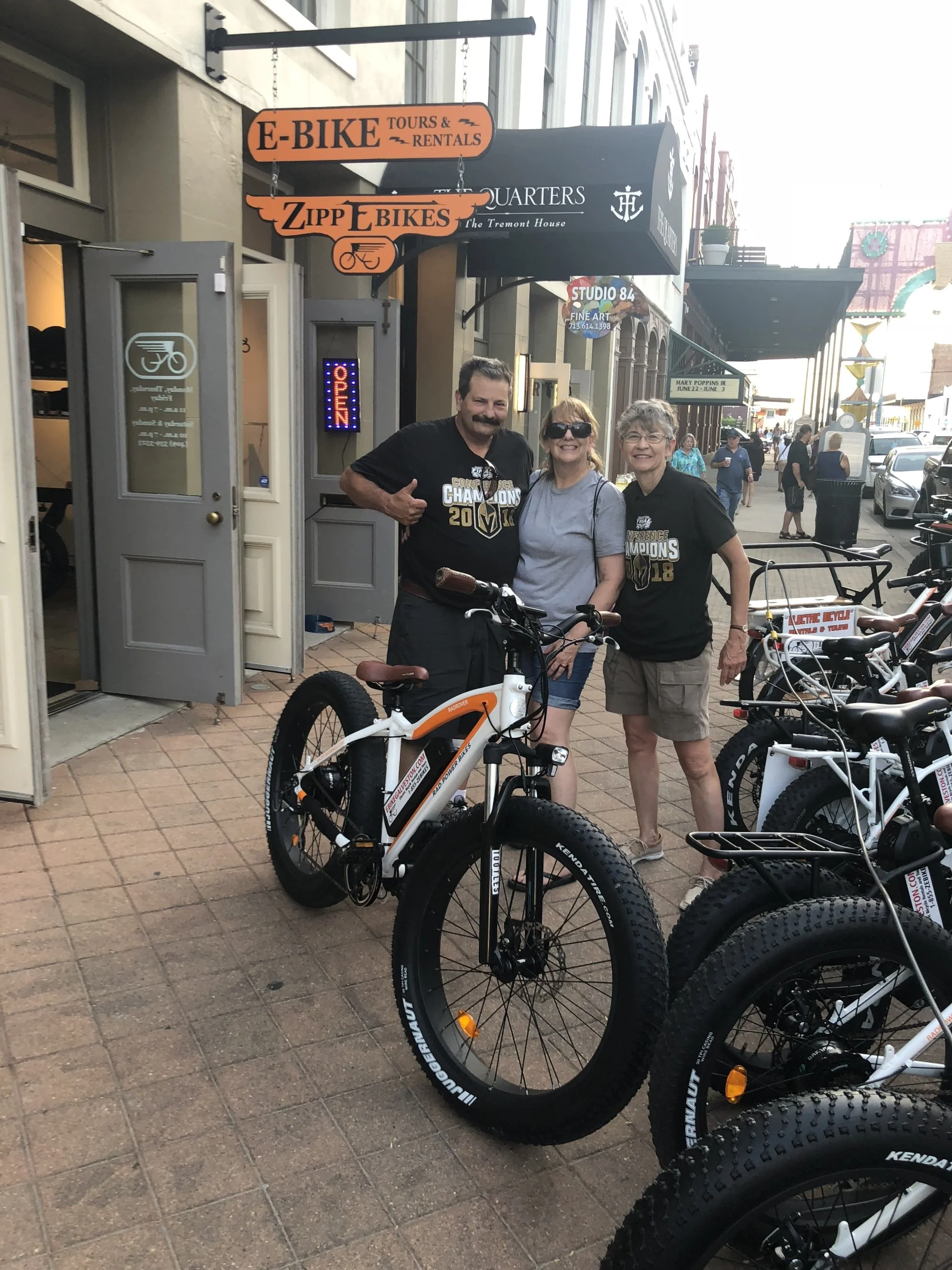 Three people standing outdoors on a brick sidewalk, smiling at the camera, with parked bicycles in the foreground and storefronts behind them.
