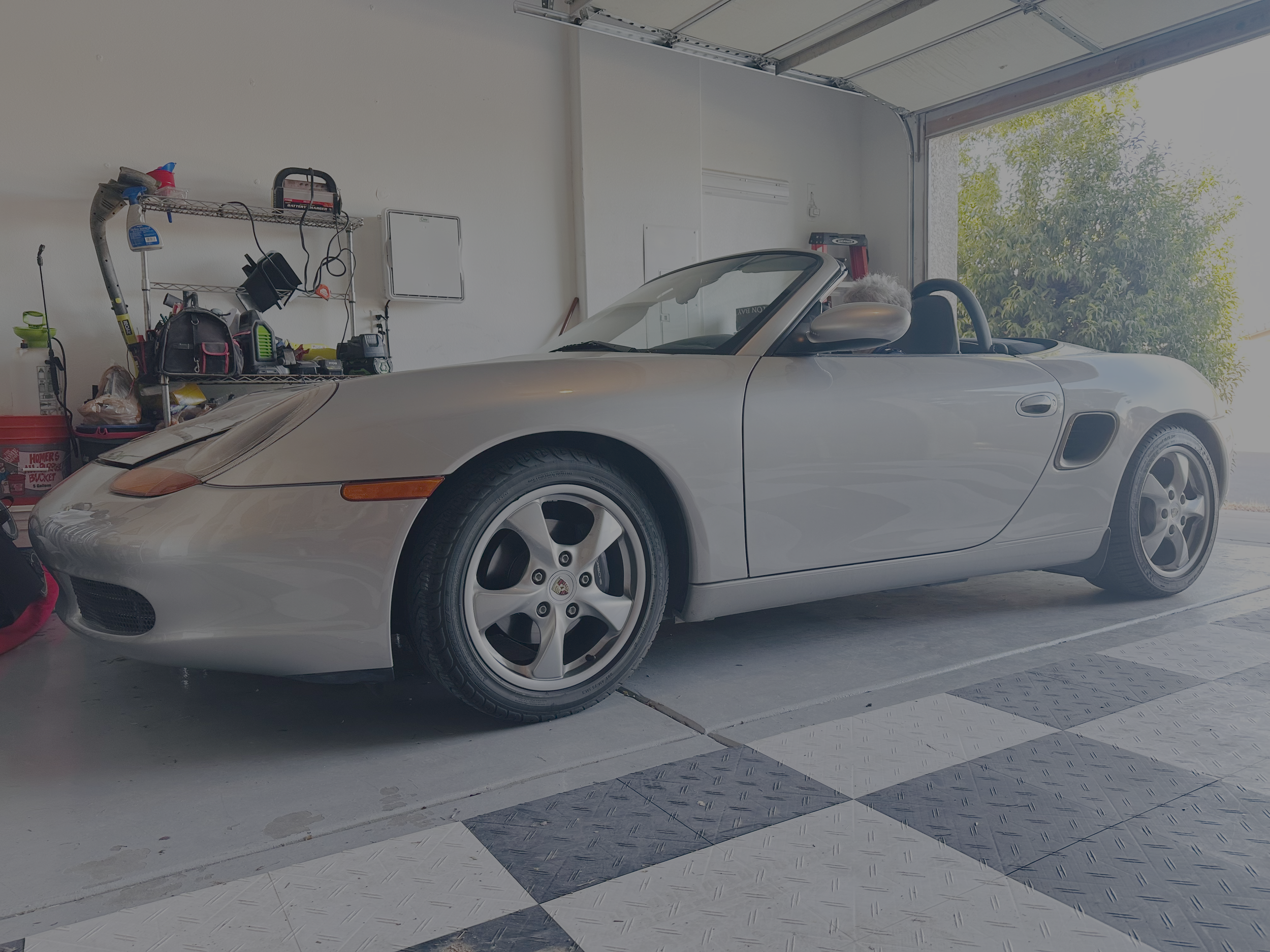 A white convertible sports car parked inside a garage with shelves and tools on the left side, and a large open garage door showing a tree outside.
