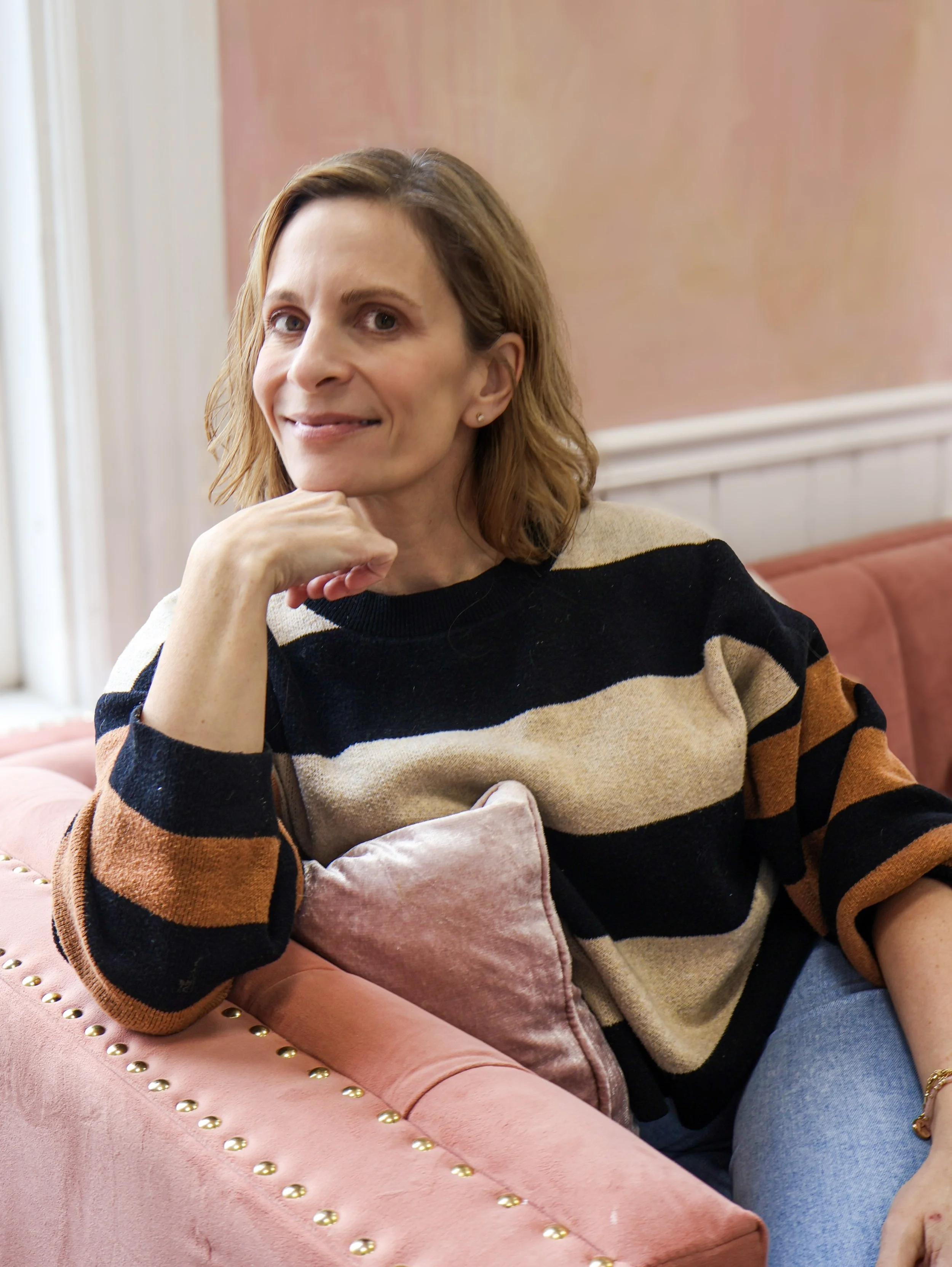 Kris Orestis, owner of Continuum Integrative Women’s Psychiatry, is sitting on a pink velvet couch with gold nailhead trim, resting her chin on her hand, with a pink pillow and a light pink wall in the background.
