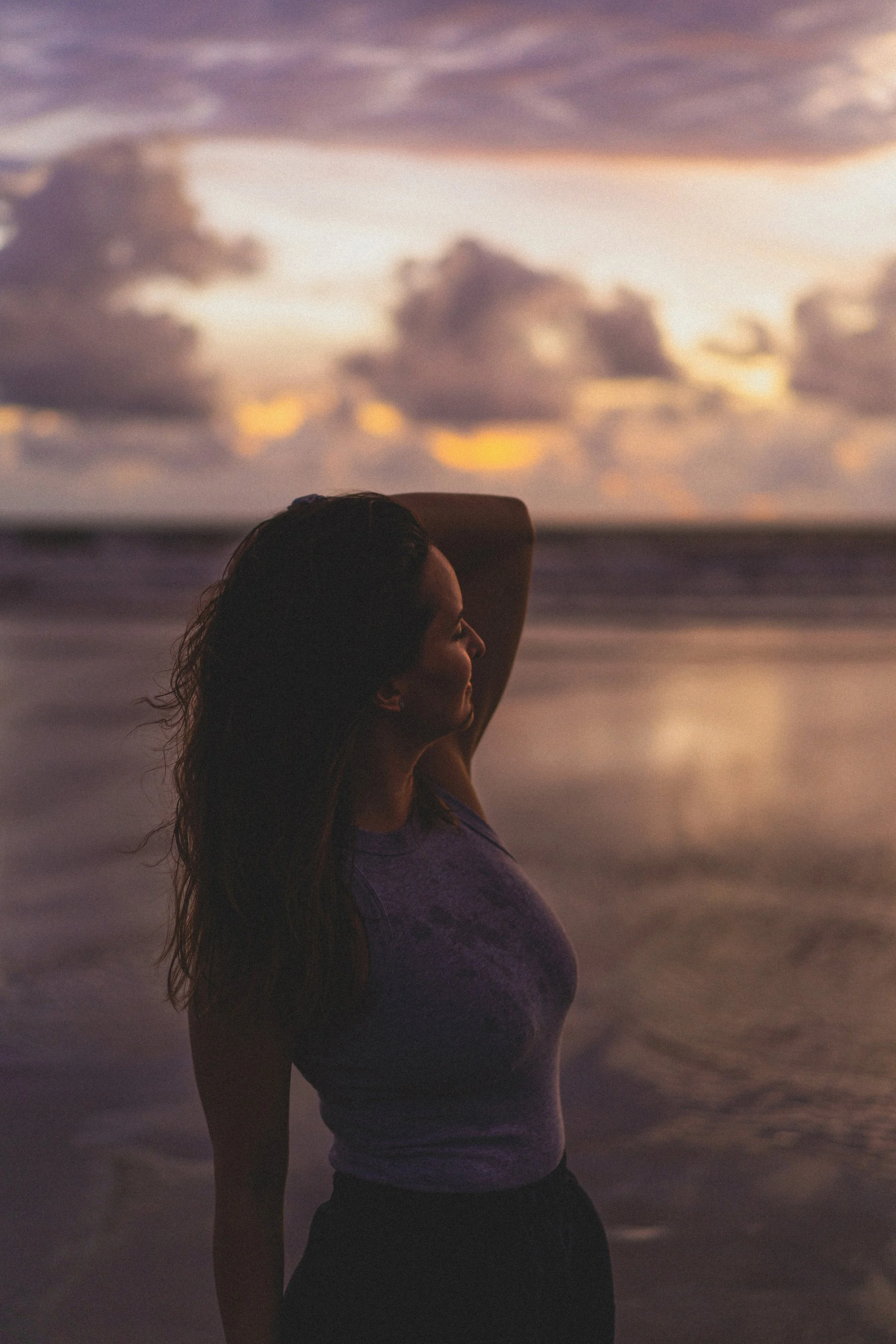 A woman standing on a beach during sunset with her arm raised behind her head, facing the ocean and sky filled with clouds.