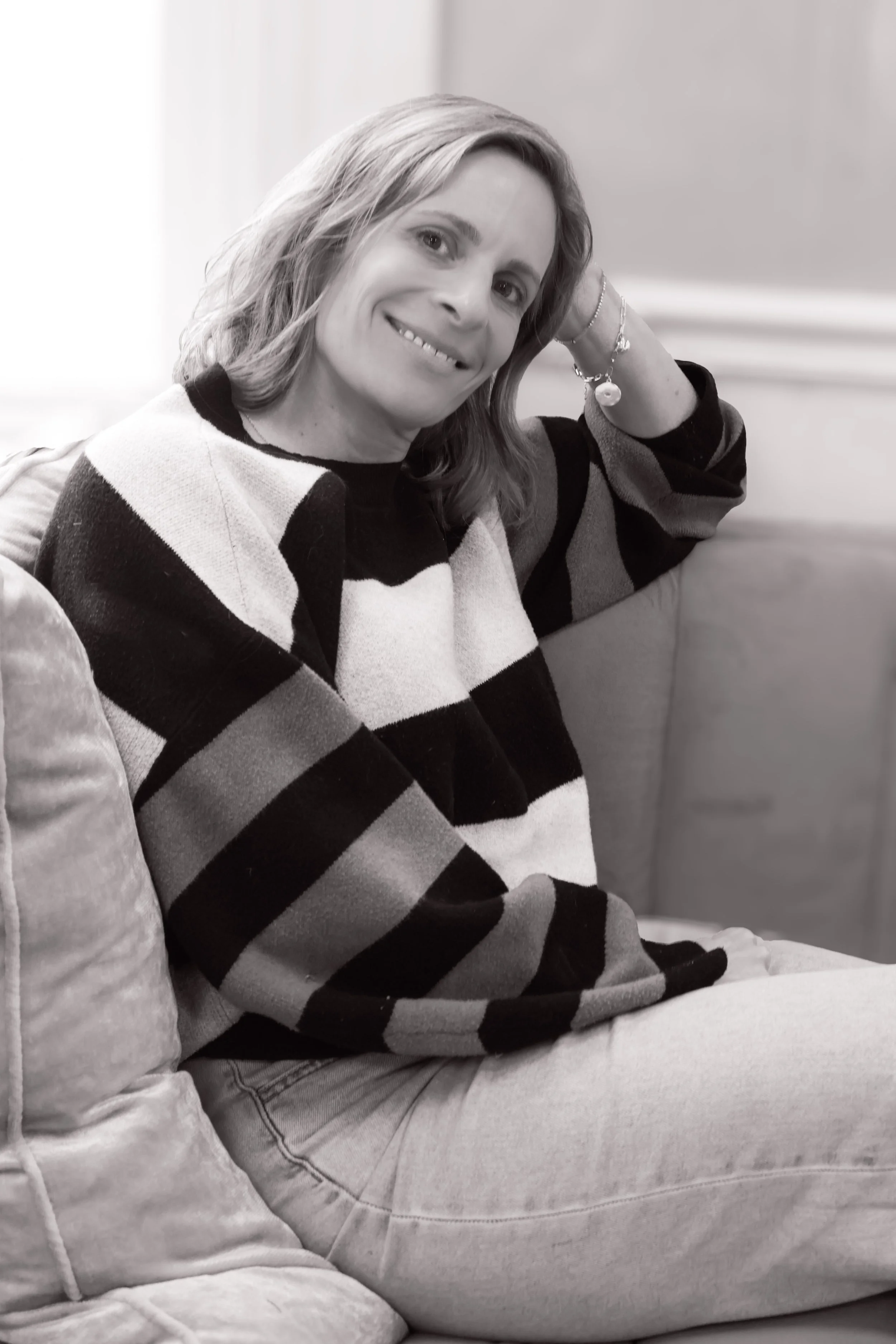 Black and white photo of Continuum Integrative Women's Psychiatric PLLC owner Kris Orestis sitting and smiling, wearing a striped sweater and light-colored pants, with one arm resting on her head.