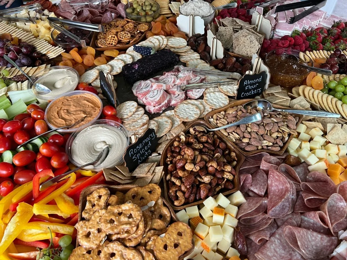 Assorted cheeses, crackers, grapes, cherry tomatoes, sliced peppers, dips, nuts, meats, and sweets on a buffet table.