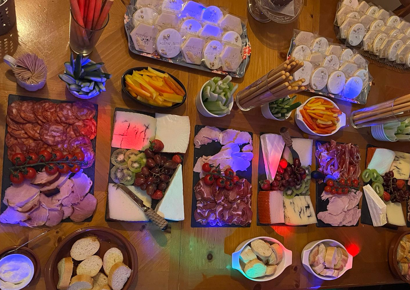 A cheese, charcuterie, and snack platter spread on a wooden table with various cheeses, meats, crackers, fruit, vegetables, and bread. There are also bowls of sliced peppers, celery, and bread slices, along with desserts or snacks in the background.