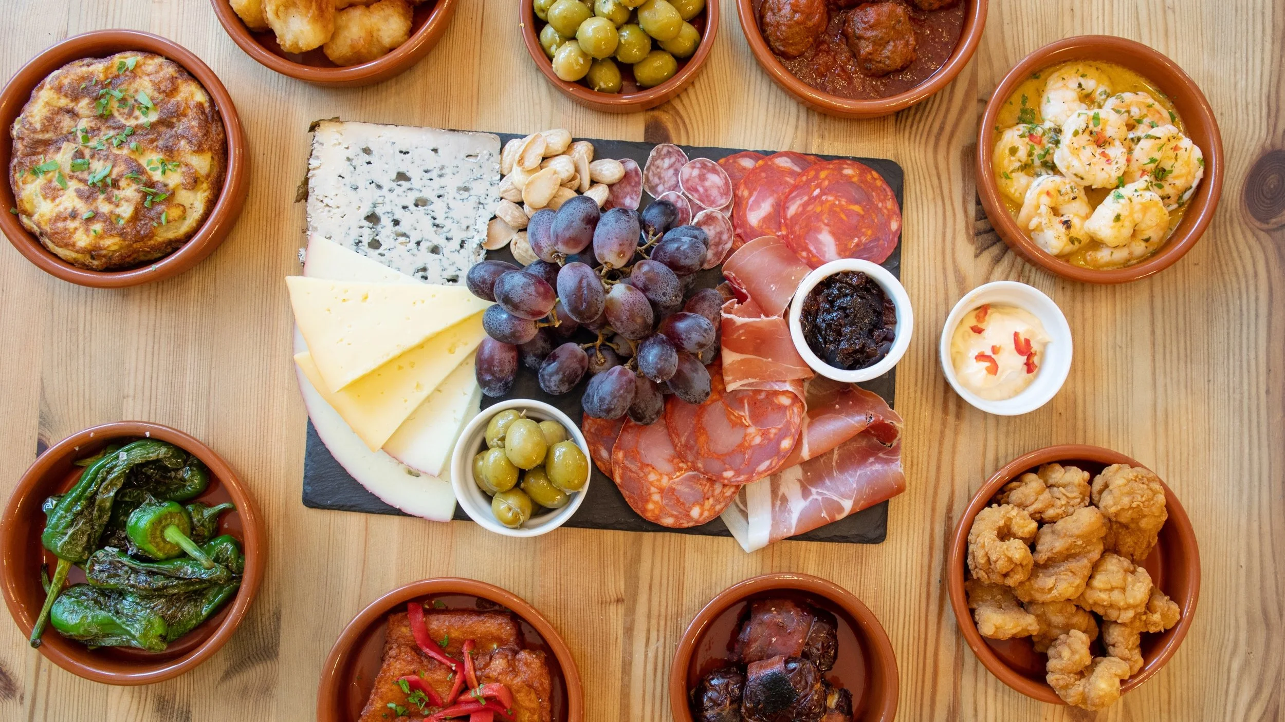A variety of Spanish tapas including cheese, grapes, cured meats, shrimp, roasted peppers, fried chicken, and other small dishes served in small bowls on a wooden table.